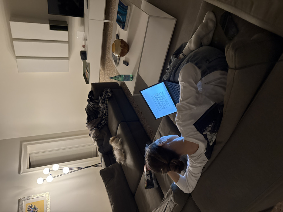 The image displays an interior living space in Venice, Italy, under artificial lighting. A female individual is situated on a dark gray L-shaped sectional sofa, her back to the viewer. She wears a white long-sleeved shirt, blue jeans, and white socks, with her hair in a bun, and is engaged with an open laptop displaying text and a table on her lap. A small, light-colored dog is curled up and resting on an adjacent section of the sofa. A pile of clothing, including a plaid shirt, is draped over another part of the sofa. In the foreground, a white rectangular coffee table holds a green glass bottle of San Pellegrino water, a brown and beige ceramic decorative item, a black remote control, and two books. The far wall features a modern white modular wall unit with closed cabinets, positioned below a visible flat-screen television on the right side. A floor lamp with five spherical white lights stands in the background near a partially open white door. The floor is a light-colored, speckled terrazzo.