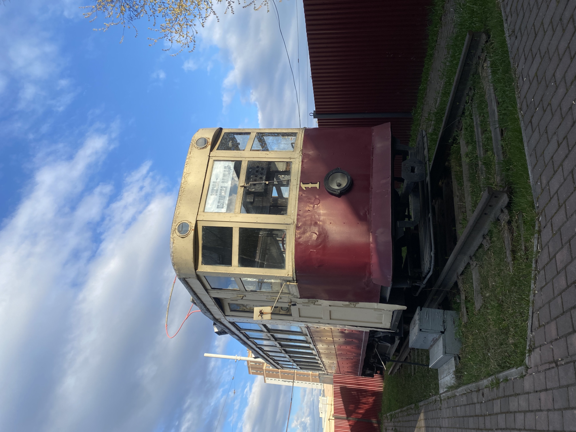 An old, two-toned tram car is prominently featured, stationary on tracks, under a clear blue sky with scattered white clouds. The tram's body is a deep maroon or dark red, while its upper section, window frames, and roof are a lighter beige or yellowed white. It features large, multi-pane windows and a circular headlight on its front, with two smaller, round lights above the windshield. The number "1" is visible on the front of the tram. Inside the front window, a destination sign displays Cyrillic text: "ВОКЗАЛ - ПЛОЩАДЬ ВОССТАНИЯ," which translates to "STATION - REVOLUTION SQUARE."
The tram is situated on grassy, overgrown tracks, suggesting it might be a display piece or retired. To the left (from the viewer's perspective, assuming the image is rotated upright), a red corrugated metal fence runs parallel to the tram. To the right, there is a paved path or sidewalk made of dark brickwork. In the upper left corner, bare tree branches with small buds extend into the sky. In the background, past the tram and fence, several modern, multi-story buildings are visible in the distance. The scene appears to be set during daytime in Yekaterinburg, Russia, with bright, natural light. No people are visible, and there is no activity taking place.