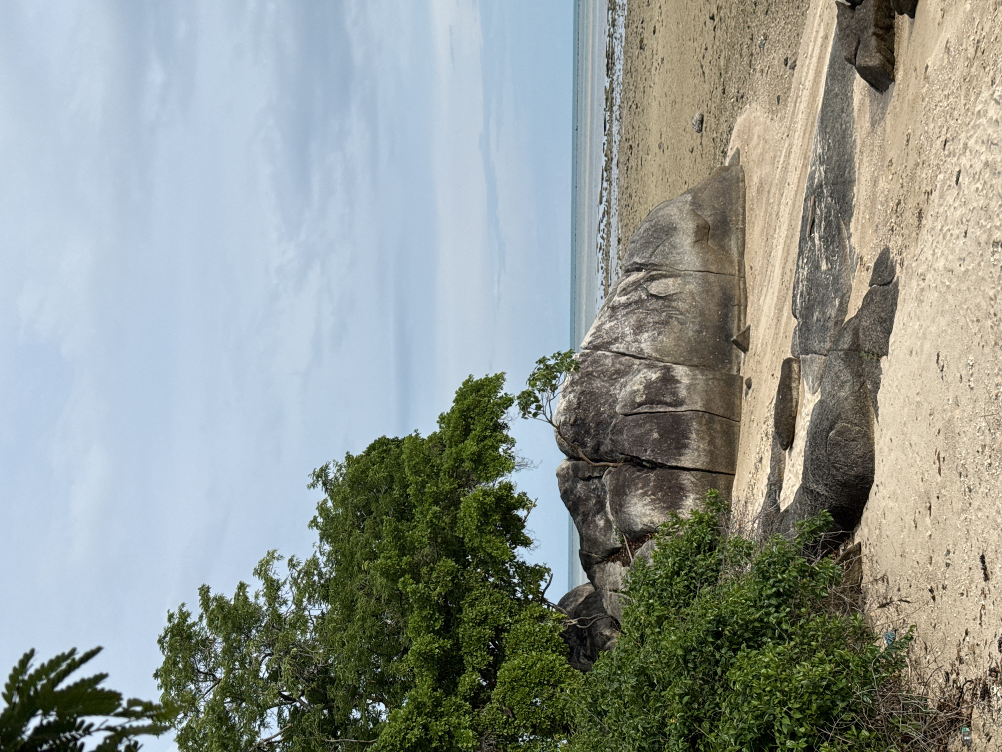 The image depicts a beach scene in Ban Bang Khao, Thailand. In the foreground, there is light-colored sand with some scattered shells and debris. Several large, dark grey boulders are visible on the sand, some with visible cracks. To the left, a lush green tree with dense foliage overhangs the scene, and a thick bush is also present. Beyond the sand and boulders, the ocean is visible, with calm, light blue water extending to the horizon. The sky is overcast with light grey clouds. There are no people or animals in the frame.