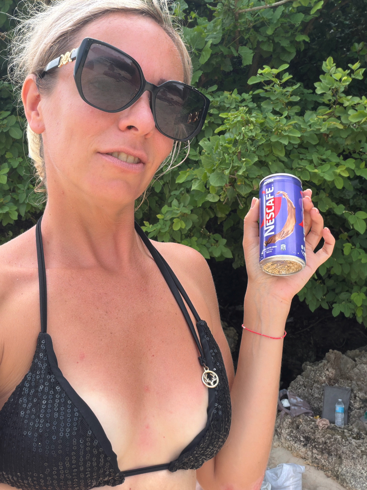 The image shows a woman holding a can of Nescafé in front of a backdrop of green foliage. She is wearing a black sequined bikini and large black sunglasses. Her hair is blonde and pulled back. The can is purple and features the Nescafé logo and an image of a chocolate swirl. The bottom of the can is filled with a granular substance. Her right hand has a thin red bracelet. In the background, to the right, there are rocks and some scattered items on the ground, including a bottle of water and a gray bag. The overall setting appears to be outdoors, possibly near a beach or natural area, consistent with locations in Malay, Philippines, such as Boracay island.