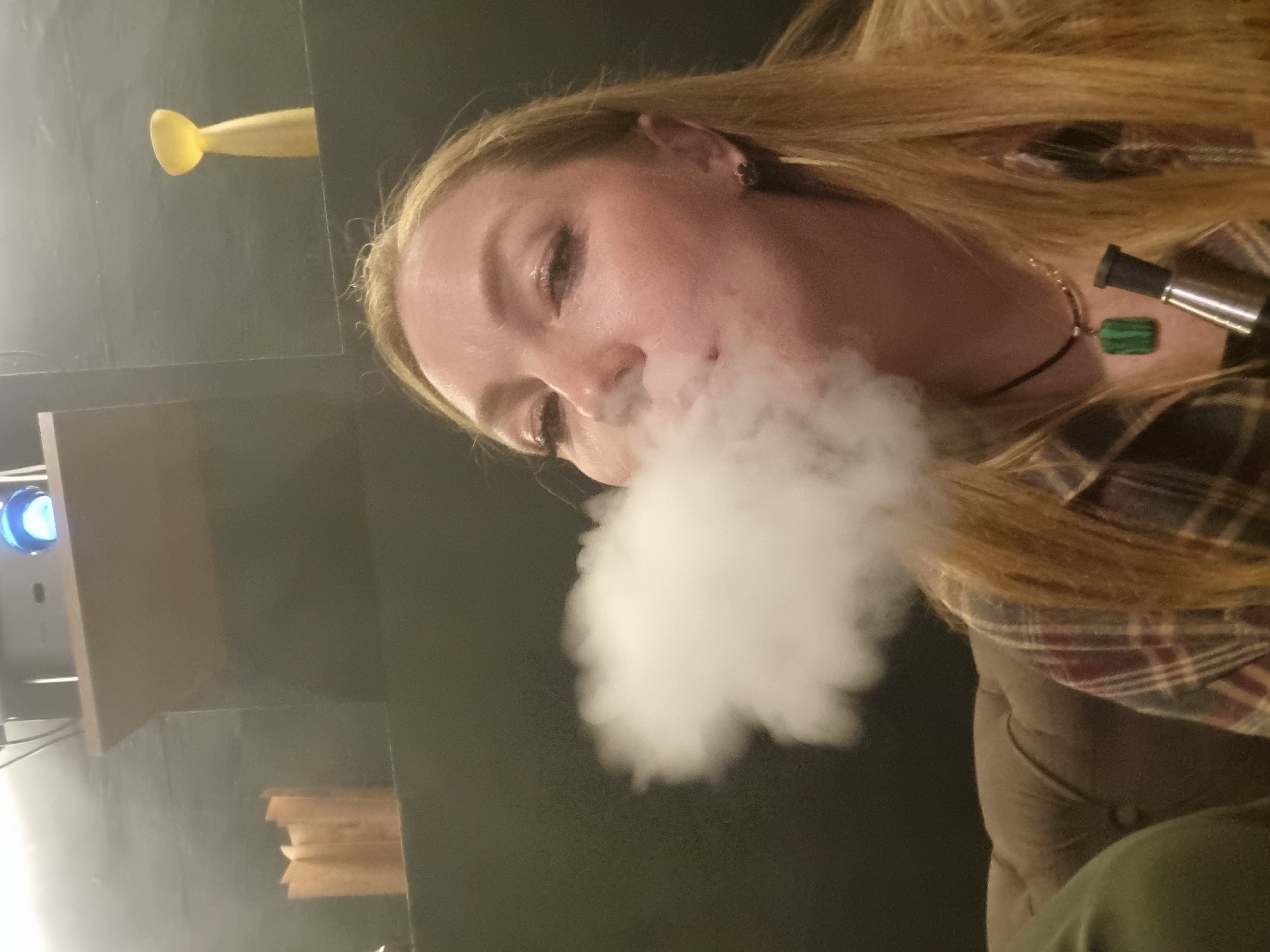 This image, taken in Chaikovsky, Russia, shows a close-up, angled view of a person with long blonde hair, exhaling a large cloud of white vapor. The person's face is visible, with eyes partially closed, suggesting focus on the act of vaping. They are wearing a plaid shirt with shades of red and brown, and a dark cord necklace with a rectangular green pendant is visible around their neck. A metallic vape device is partially seen near their right shoulder.
The setting appears to be indoors, likely in the evening or at night, given the dim, artificial lighting. In the background, to the left of the person's head, there's a dark surface from which a distinct yellow, elongated object protrudes. Further to the left, a light source or projector emitting a blue glow is visible on a shelf or surface. The person seems to be seated on an upholstered chair or sofa, indicated by the visible fabric texture and folds. The overall mood is relaxed and casual. There is no visible text in the image.