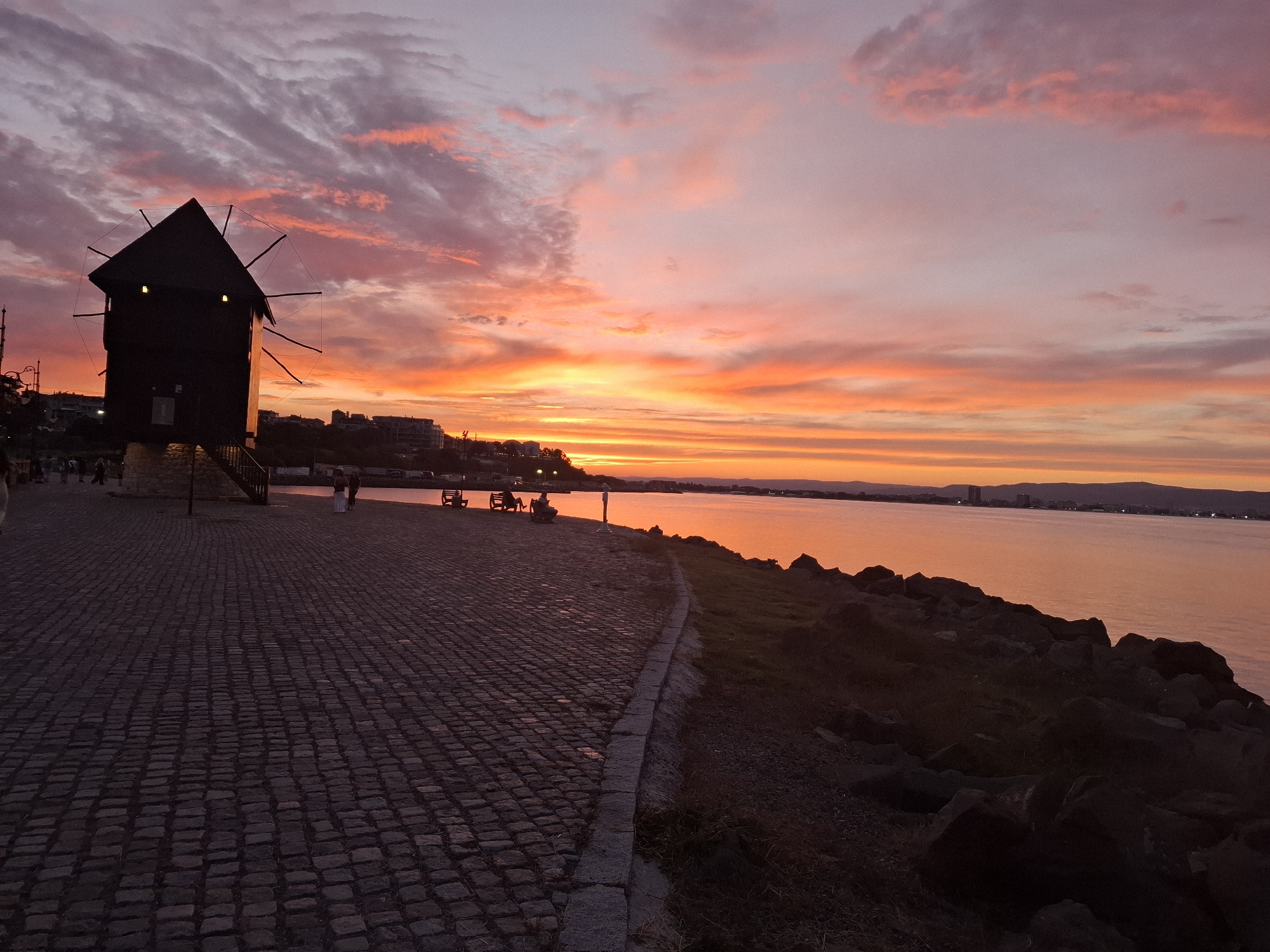 The image depicts a picturesque sunset over the Black Sea in Nesebar, Bulgaria. The sky is a vibrant canvas of pink, orange, and yellow hues, with dramatic clouds creating a sense of depth and movement. The sun, not directly visible, is low on the horizon, casting a warm glow across the water.
In the foreground, a cobblestone pathway leads the viewer's eye towards the water. To the left, silhouetted against the colorful sky, stands a dark, wooden windmill, a characteristic landmark of Nesebar. Its iconic structure, with a pointed roof and visible blades, adds a historical and rustic element to the scene. Several people are visible near the windmill and along the path, some seated on benches, enjoying the view. The path itself is made of rough, uneven stones, indicating an older, possibly historic, area.
The scene transitions to a rocky shoreline bordering the sea. The water is calm, reflecting the intense colors of the sunset. In the distance, the silhouette of a town or city can be seen along the coastline, with buildings and lights hinting at urban life. The overall atmosphere is serene and tranquil, evoking a sense of peace and wonder inspired by the natural beauty of the sunset. The time of day is clearly sunset, with the fading light creating dramatic shadows and illuminating the sky. The weather appears clear, allowing for the unobstructed display of the sunset's spectacle. The presence of the windmill and the distant cityscape suggest a coastal town or historical area.
