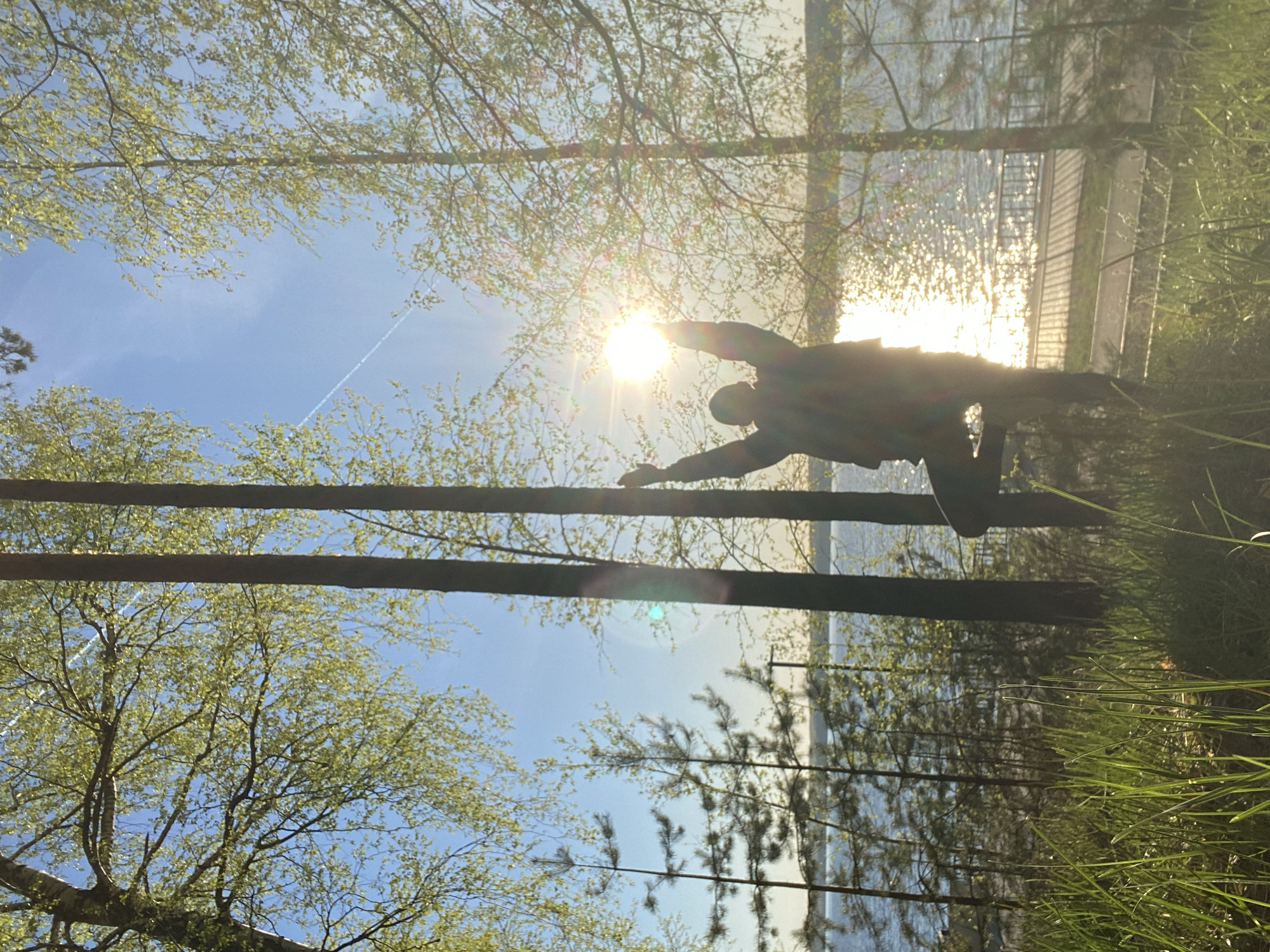 The image, apparently taken from a low angle, depicts a sunlit outdoor scene. A person silhouetted against the bright sun stretches their arms out, appearing to touch or hold onto two horizontal wooden poles. Behind the person, there is water and a building. Trees are visible in the foreground and background, some with green foliage. The sky is blue. The location is Chaikovsky, Russia.