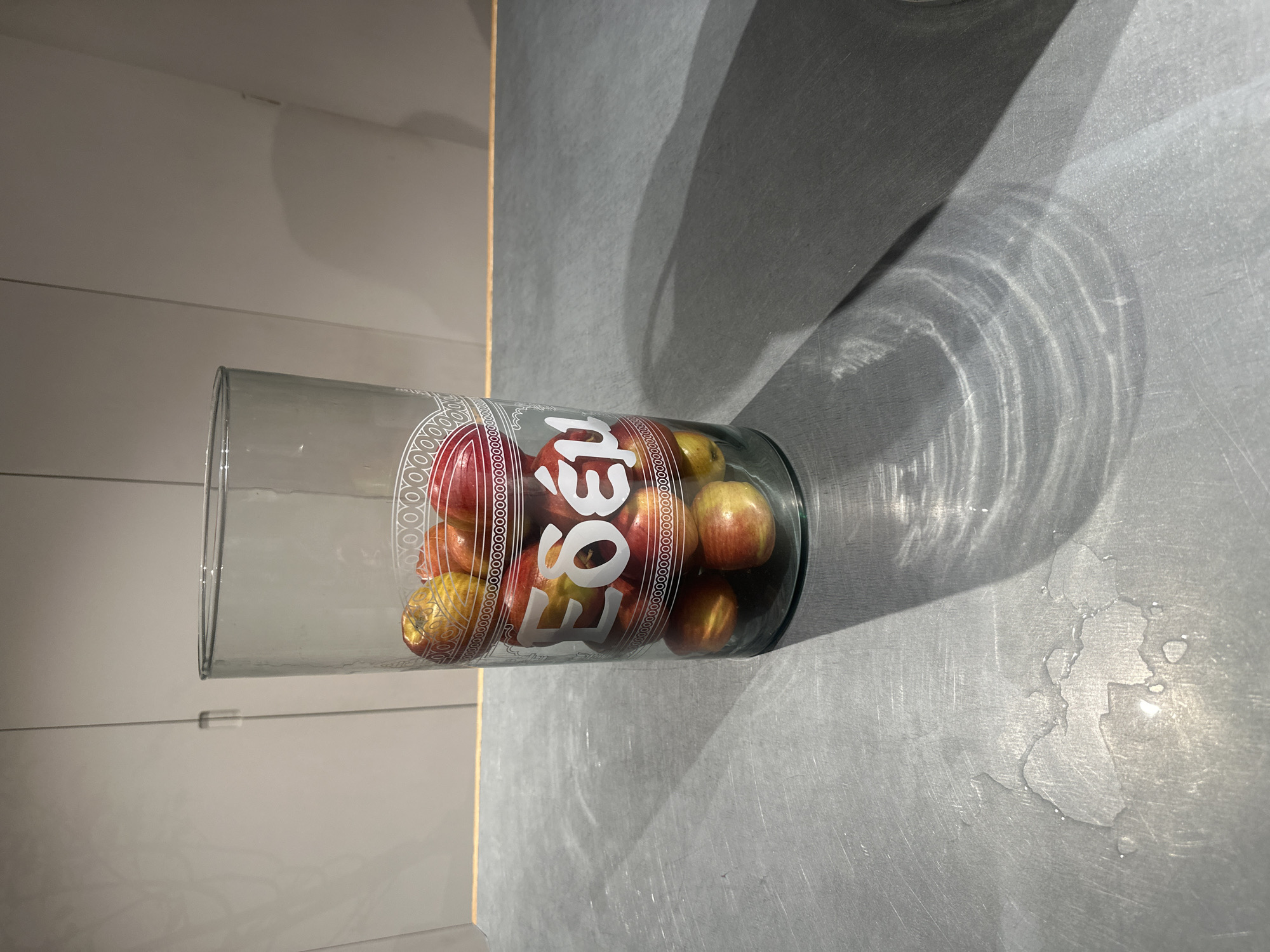 A vertically oriented image captures a clear glass cylinder or vase, placed horizontally on a dark grey, slightly reflective surface. The glass is filled with small, mostly red and yellow apples. To the left of the glass, a light grey wall or cabinet with subtle horizontal paneling is visible, separated from the grey surface by a thin vertical golden line.
Strong directional light, originating from the right, casts prominent shadows of the glass and its contents onto the grey surface. This light also creates intricate patterns of refracted light (caustics) on the surface, indicative of light passing through the glass and the apples. Several water droplets or a wet area are visible on the grey surface in the foreground, glistening under the light.
On the side of the glass, white, stylized text is clearly visible, reading "EGE" followed by "1393". This text is flanked by decorative bands of small, interconnected circles. The apples inside appear firm and vary slightly in their red and yellow hues. The scene, likely in a domestic kitchen setting in London, United Kingdom, is illuminated by natural light, suggesting it is daytime.