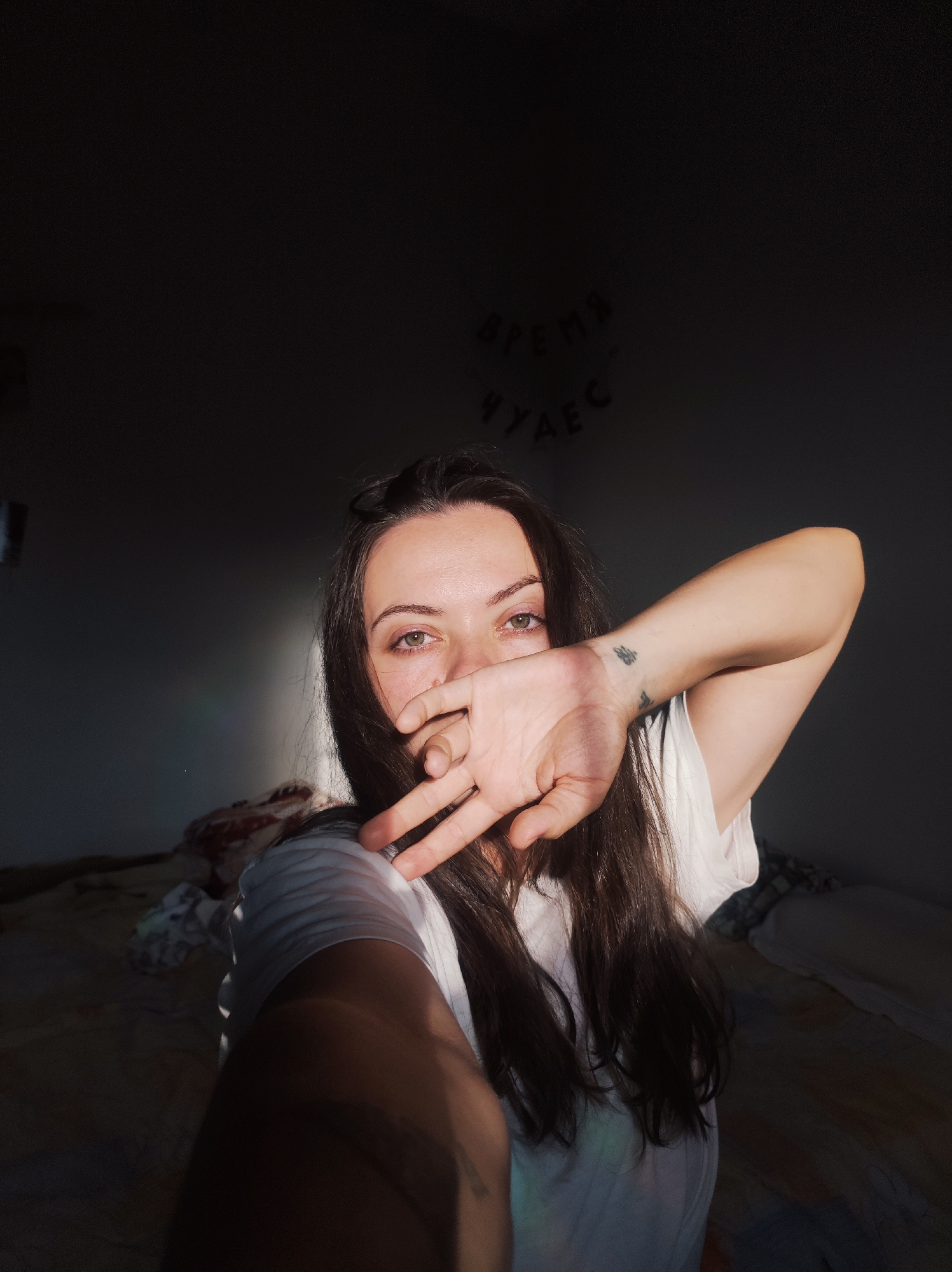 A close-up selfie features a young adult female, partially obscuring her face with her left hand, in an indoor room in Unknown, Unknown. She looks directly at the camera with green/hazel eyes. Her left hand, covering her mouth and chin, displays a small, dark, abstract tattoo on the wrist. Her dark, long hair frames her face, and she wears a white t-shirt. Strong, warm, directional light, likely sunlight, enters from the left, illuminating her left side and casting deep shadows across the right side of the image and background. A faint rainbow-like lens flare is visible on the left. The dark wall in the background displays decorative, irregularly placed dark Cyrillic letters that spell "ВРЕМЯ ЧУДЕС" (VREMYA CHUDES), translating to "TIME OF MIRACLES." A bed with dark bedding and other draped fabrics is partially visible in the shadowed background on both sides.