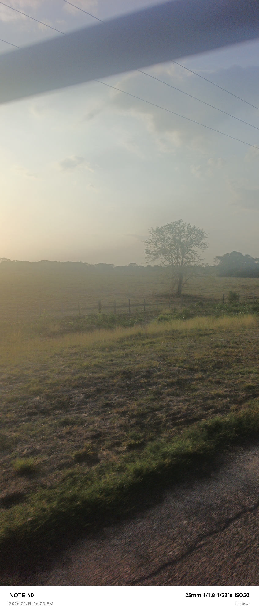 Here is a detailed description of the image:
**General Description:**
The image captures a misty, rural scene, likely taken at dawn or dusk, characterized by a haze that obscures distant features. A large shadow from a man made object dominates the sky.
**Environment and Landscape:**
The landscape is dominated by a large field that transitions from dry in the foreground to green further back, suggesting varying soil moisture levels. A solitary, leafy tree is positioned at the center of the midground, while hints of denser vegetation, likely trees and shrubs, line the horizon. There is a wooden fence present.
**Atmospheric Conditions:**
The heavy mist obscures the background, creating a soft, ethereal quality. The sun is behind the observer, making the scene hazy.
**Objects and Elements:**
A large shadow runs across the sky.
**Scene Location and Time:**
Based on the OCR text, the location is specified as "El Baul," and the time and date are indicated as "2026.04.19 06:05 PM." It is important to note that the reference to La Cruz, Venezuela, is not mentioned in the original data provided; therefore, I can only describe the scene as it appears based on the data and image.
**Overall Impression:**
The image is a pastoral scene that relies on atmospheric conditions to create a sense of calm and mystery.