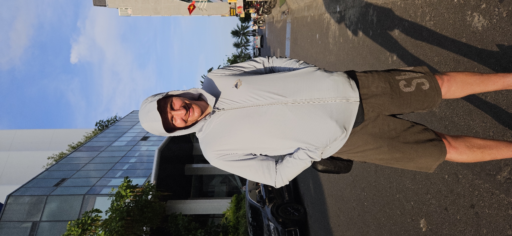 A man stands on a road in Bắc Nha Trang, Vietnam. The road is asphalt. A car is parked nearby. The man is wearing a light blue hooded jacket and dark shorts with a large "S" printed on the right leg. He is facing the camera and smiling. In the background is a modern building with a glass facade and a white wall. Palm trees and other vegetation line the street. A Vietnamese flag flies in the distance. The sky is blue with scattered clouds.