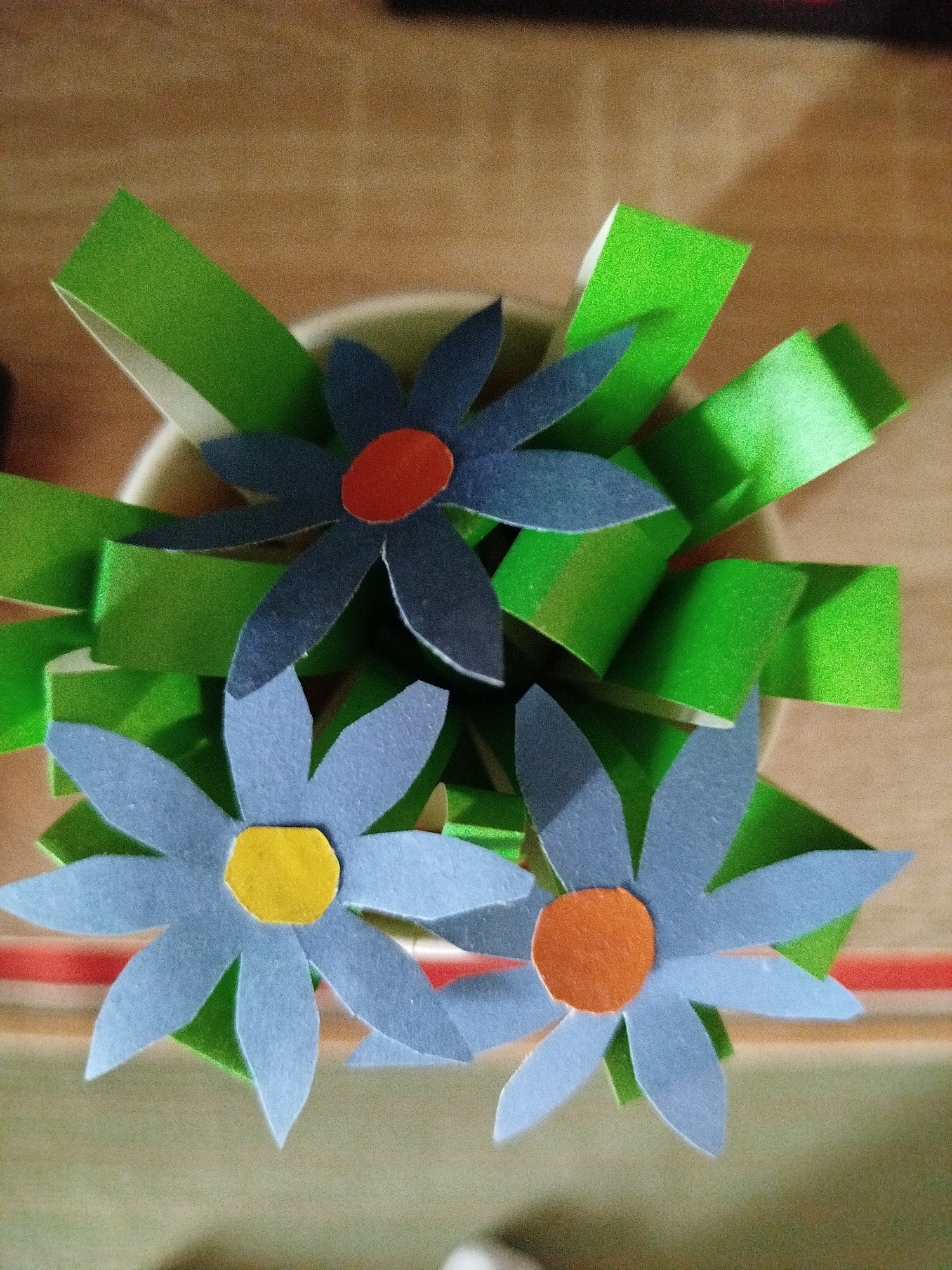This is a close-up, top-down, slightly angled shot of a handmade craft bouquet of paper flowers and green paper leaves. The bouquet is arranged in a white cylindrical container which is partially visible. Three blue paper flowers are prominently displayed, each with a different colored center: one has an orange center, one has a yellow center, and the third flower is partially obscured but appears to also have an orange center. The green paper leaves are cut into strips and appear to be folded or curved, giving them a three-dimensional effect. The background is a textured, light brown surface, possibly a wooden table. The overall lighting is somewhat dim, with some areas of highlight on the glossy paper of the green leaves. There are no people or animals visible in the image, and the scene appears to be indoors. The geographical location of Крывасельскі сельскі Савет, Belarus is not discernible from the visual information provided in the image.