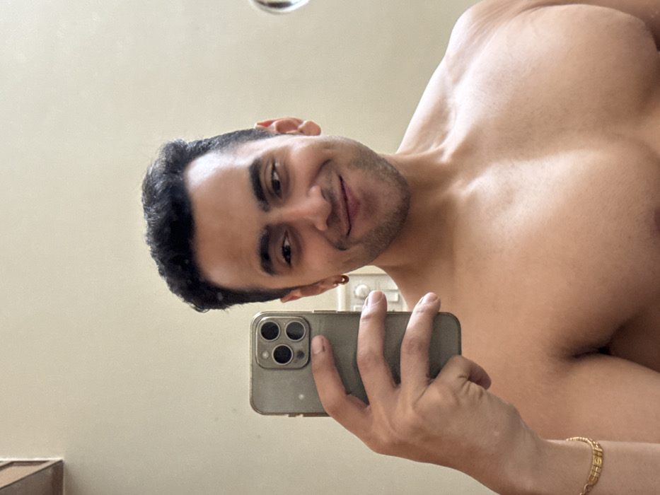 A shirtless man takes a selfie with his phone in a bathroom. He has dark hair and a slight stubble, and he is smiling. The phone case is gray and covers the camera. His chest and shoulders are muscular. A gold bracelet is visible on his wrist. The background is a plain, light-colored wall. An earring is seen in his ear.