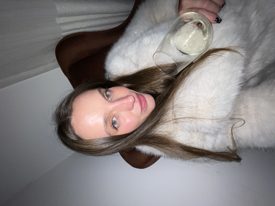 A woman with long brown hair poses indoors. She wears a fluffy white fur coat and sits in a brown chair. She holds a glass of champagne. She has fair skin, pink lips, and light-colored eyes. The background consists of a white wall and the edge of window blinds. The photo appears to be taken from a low angle and rotated counter-clockwise.