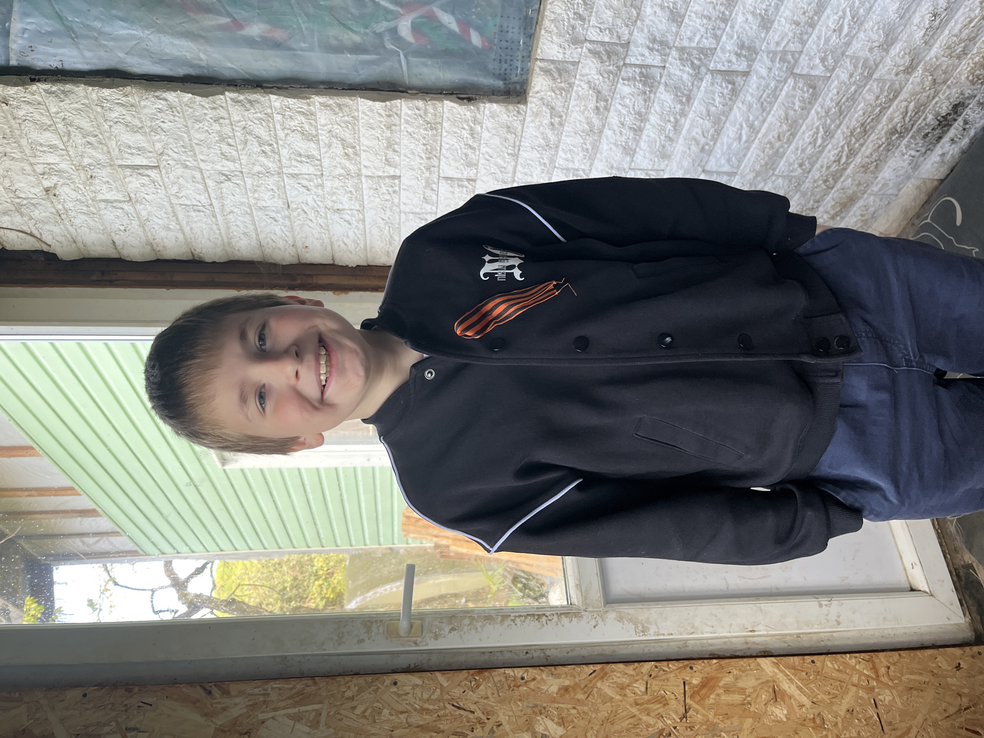 A young boy with short brown hair and a cheerful expression poses for a photo indoors, in what appears to be Vesyoloye, Russia. He is smiling broadly and looking directly at the camera. He wears a dark-colored (likely black) jacket with white piping along the shoulders and sleeves, and several dark snap buttons down the front. Pinned to the left chest of his jacket is a distinct orange and black striped St. George's ribbon. Above the ribbon, a white emblem or logo is visible on the jacket. He is also wearing dark blue pants.
The boy is positioned in front of a window. Through the window to his left, a light green exterior wall or siding is visible, along with some trees and foliage, indicating daylight outside. To his right, an interior wall with a textured white, brick-like or stone-like finish is seen. Above this textured wall, a reflective surface, possibly a window, shows a blurred red and white striped pattern. The flooring visible in the foreground and beneath the window frame is made of oriented strand board (OSB) or particleboard. The window itself has a white frame. The overall scene suggests a casual indoor setting, illuminated by natural light. The boy's happy demeanor is the prominent emotion conveyed.