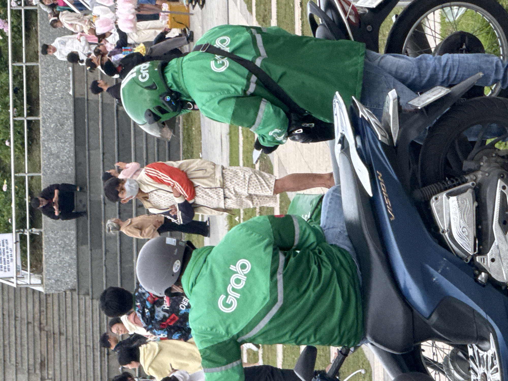This image depicts an outdoor scene in Da Lat, Vietnam, likely on a public staircase. Two individuals wearing green Grab driver jackets are prominent in the foreground, one on a dark blue motorcycle and the other standing. The motorcycle driver, seen from the back, wears a grey helmet and blue jeans. The second Grab driver, also viewed from behind, has a green helmet and is holding a mobile device. In the midground, several other people are visible on the stone steps, including a man in black sitting on the steps, a woman in a tan jacket, and others in casual attire. Some individuals are wearing masks. Further up the steps, a group of people, some of whom appear to be tourists, are gathered. The environment includes grassy areas and a railing with pink flowers. The overall atmosphere suggests a busy public space.