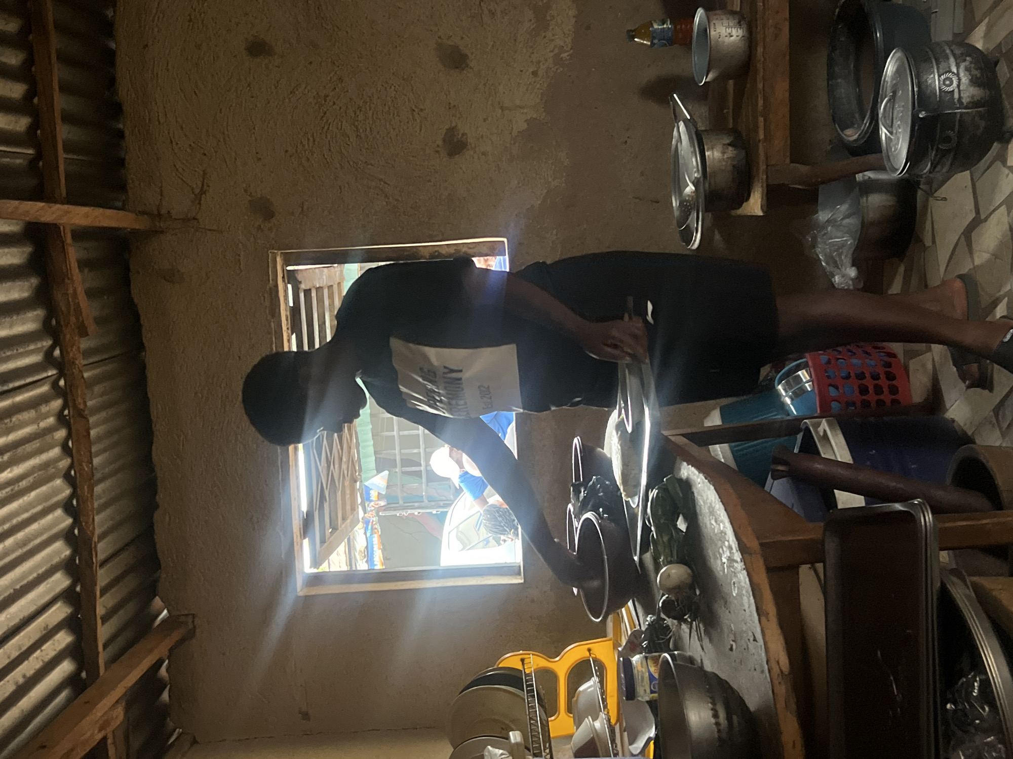 The image depicts an indoor scene, likely a kitchen or utility area, in Jos, Nigeria, where a person is engaged in an activity. The central subject is a young person, seen from behind, wearing a black t-shirt with a white graphic print and black shorts. They are bent over a wooden table or counter, actively washing or preparing something in a bowl or basin, with their hands submerged.
The room has a rustic appearance. The left wall and ceiling feature corrugated metal sheeting supported by raw wooden beams. The other visible wall, on the right, is made of rough, textured material, possibly mud or unplastered cement. A window is positioned behind the person, allowing bright daylight to stream in and illuminating parts of the scene. Through the window, other individuals are faintly visible outside, suggesting an outdoor area like a courtyard or street.
The wooden table in the foreground is cluttered with various kitchen items, including metal pots, pans, a measuring cup, ceramic bowls, a dark plastic bottle, and some fresh vegetables like onions or garlic. Adjacent to the table, shelves made of wood hold more cookware, including large pots and lids. The floor exhibits a patterned surface, possibly tiles or linoleum. A red plastic laundry basket and other containers are also visible on the right side of the image.
The bright light from the window indicates it is daytime. On the back of the person's t-shirt, text is partially visible, appearing to say "CELEBRATING" and "202" as part of a larger, unreadable phrase. The overall setting suggests a functional, lived-in space with practical amenities.