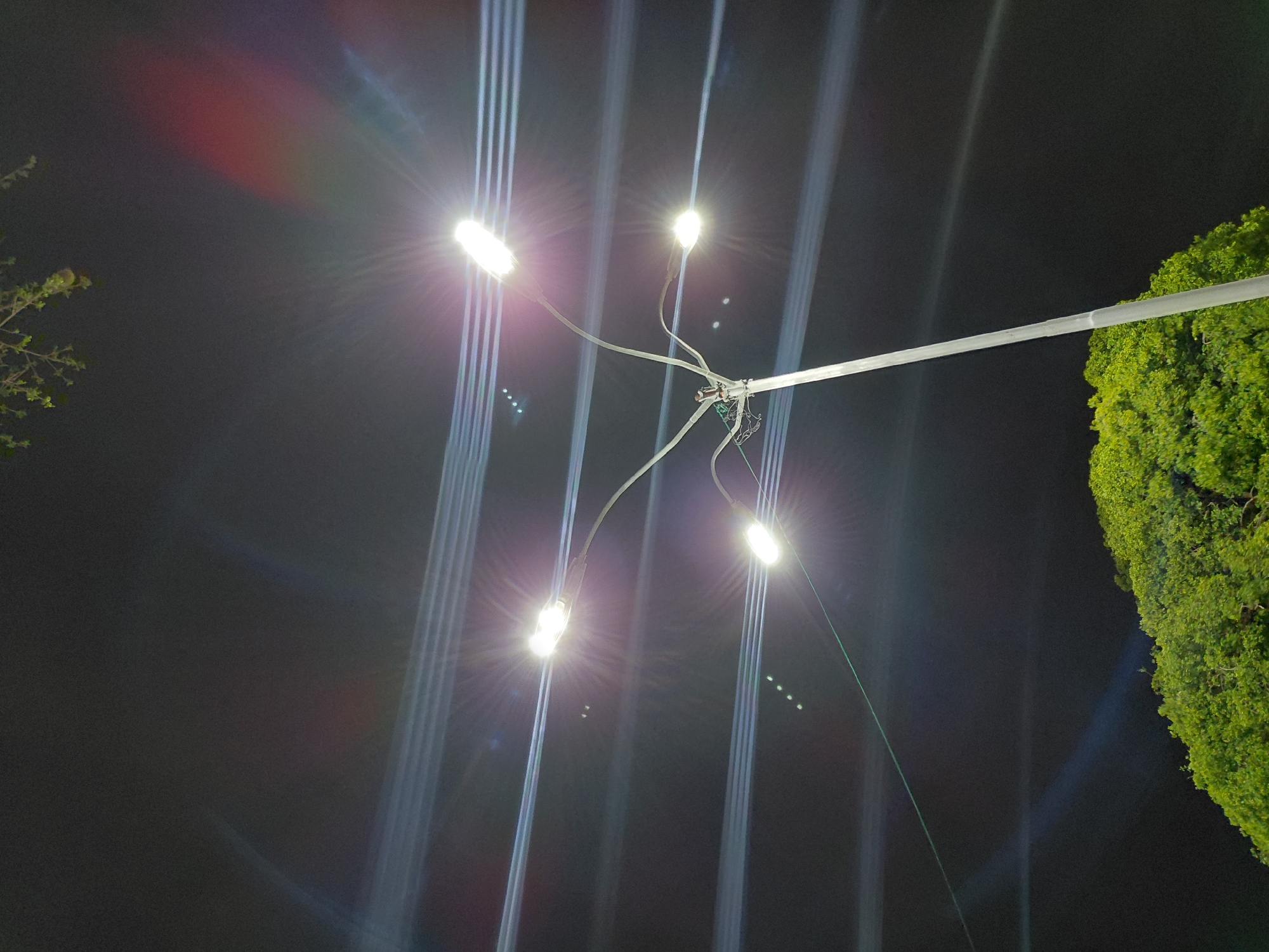 The image depicts a nighttime scene in Santa Ana, Venezuela, centered on a prominent street light structure. A tall, grey metallic pole rises from the lower-center to the mid-right, supporting a branching fixture with at least four arms. Each arm terminates in a bright, rectangular LED street light, emitting intense white illumination. These lights produce distinct starburst lens flares that radiate outwards.
The dark, nocturnal sky serves as a backdrop, displaying various optical artifacts. Horizontal streaks of light traverse the upper and middle sections of the frame, originating from the intense light sources. A noticeable reddish-purple chromatic aberration manifests as an arc in the upper right quadrant. Multiple series of small, bright, blue-green, elliptical ghosting dots are visible in curved patterns, particularly to the left of the upper lights and below the lower-left light. A faint, diagonal green line, consistent with a lens flare, extends from one of the upper light sources.
The lower portion of the image is occupied by dense, vibrant green foliage, indicative of a large tree canopy or thick shrubbery. Additional sparse green leaves and branches are visible in the top-left corner against the dark sky. The scene is devoid of any human presence, animals, or vehicles.