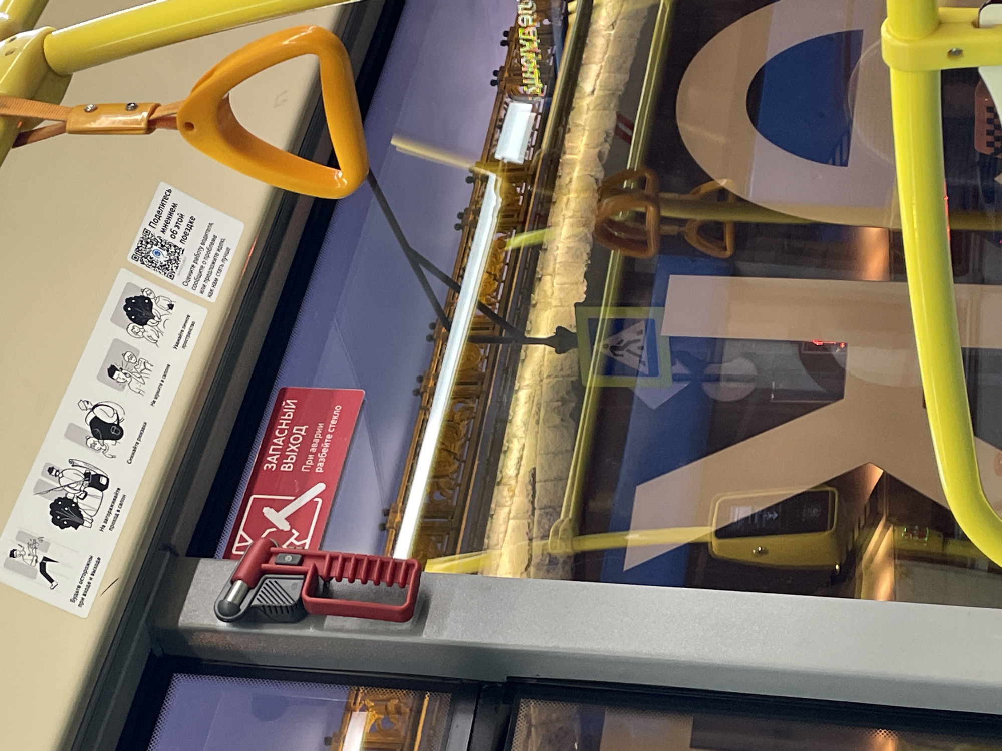 The image is taken from inside a public transportation vehicle, likely a bus or a tram, in the city of Moscow, Russia. The focus is on the safety features and informational stickers inside the vehicle. There is a yellow overhead handrail with an orange handle to the left of the frame. A sticker provides information about providing feedback regarding the ride. Additionally, there is a sticker depicting the proper etiquette for entering and riding on the bus (removing backpacks, not blocking the aisle). A red sign indicates the emergency exit with an adjacent tool to break the glass, should there be an emergency. In the reflection on the window, a street view can be observed, including what appears to be a pedestrian crossing sign and street lights.