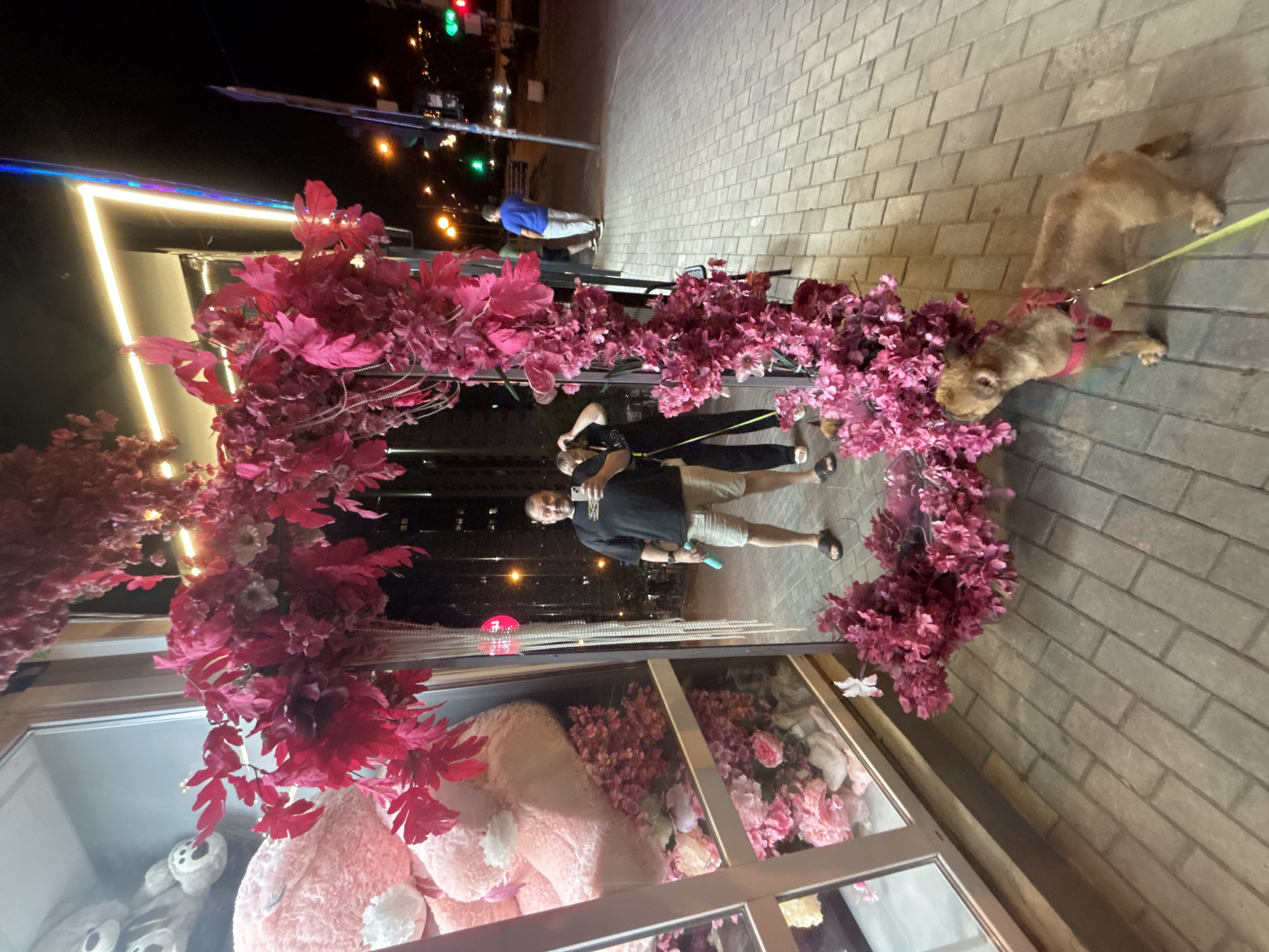 In Krasnodar, Russia, a man and a woman are standing in front of a storefront decorated with a heart-shaped floral arrangement. The man, holding a smartphone, is taking a selfie, while the woman stands beside him. A small, light-brown dog on a leash is also present in the foreground. The storefront displays large stuffed teddy bears through its window. The scene is on a paved sidewalk, with a street and traffic lights visible in the background, suggesting an urban nighttime setting.