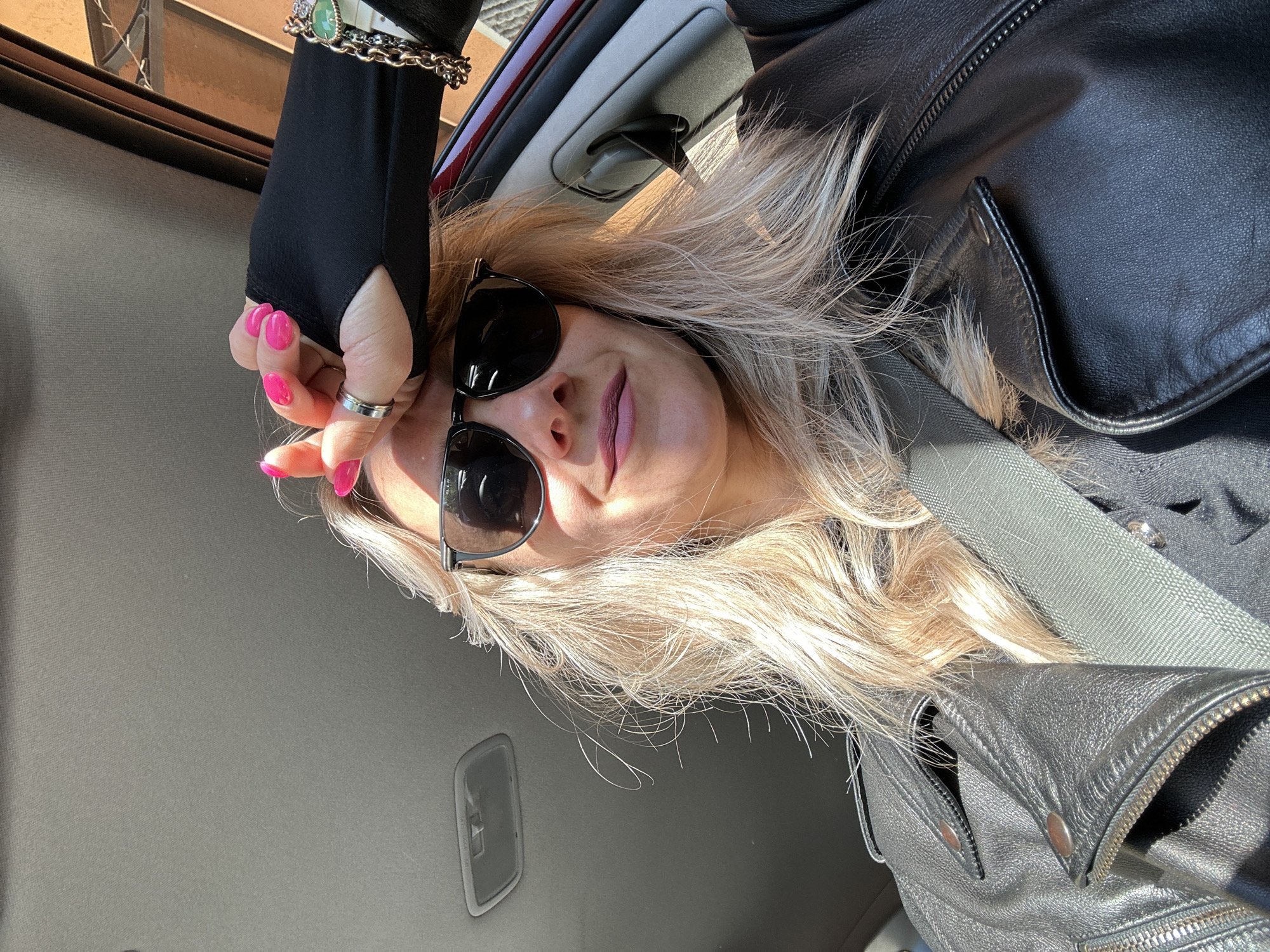 A person is in a car, wearing a black leather jacket and sunglasses. The person has blond hair and pink nail polish. The person is also wearing a black glove with a ring and a bracelet. The car's interior is visible. The image appears to be taken from inside the car. There is no definitive information from this image to say that the location is Sesto Campano, Italy.