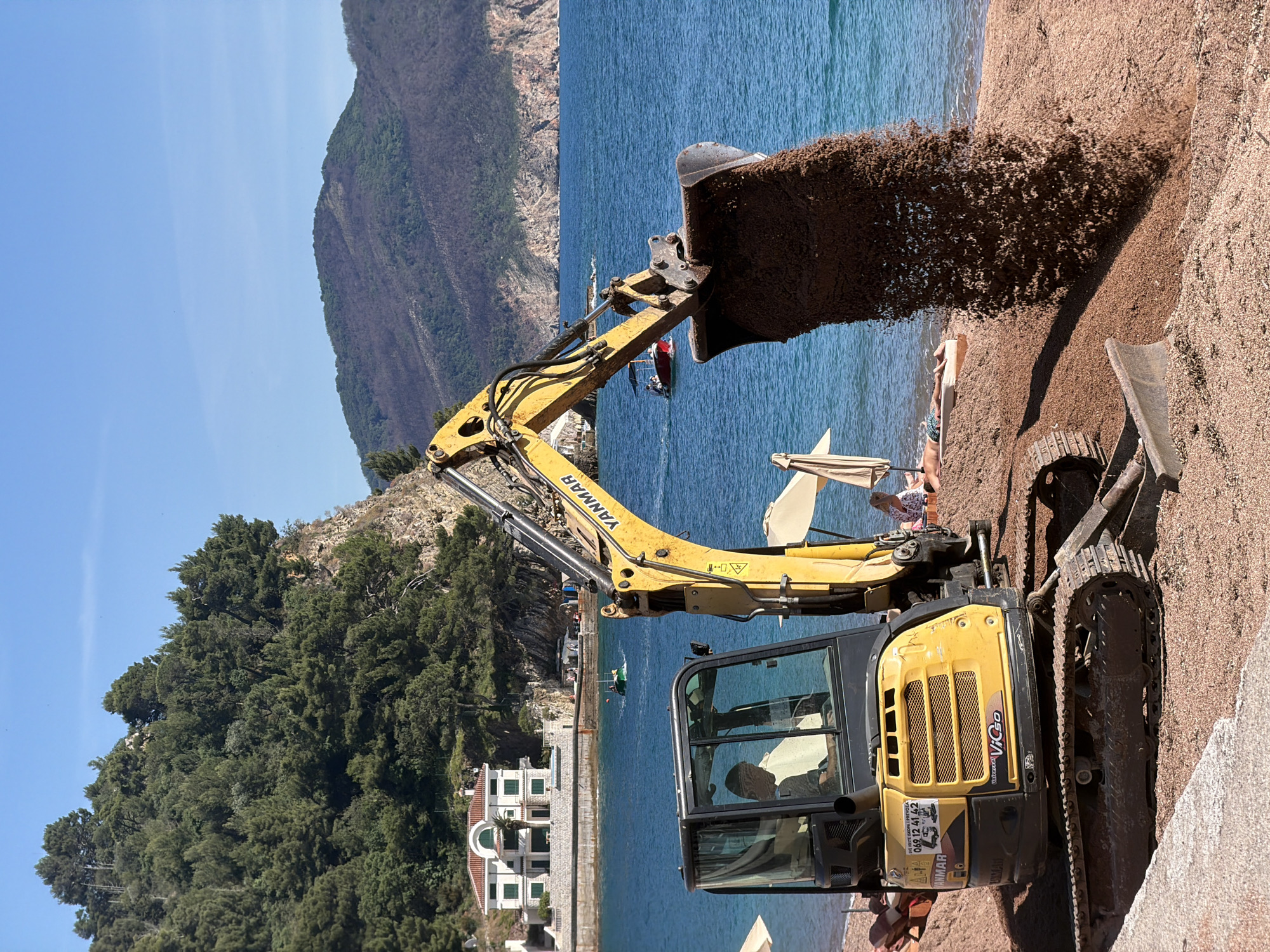 A yellow Yanmar excavator is dumping sand onto a beach in Petrovac centar, Montenegro. The excavator is positioned on the beach, with the arm extending over the sand and the bucket dumping the sand. There are people lounging on the beach near the excavator, and a sun umbrella is nearby. Behind the beach is the Adriatic Sea, with a mountain in the background. A building is visible on the coast.