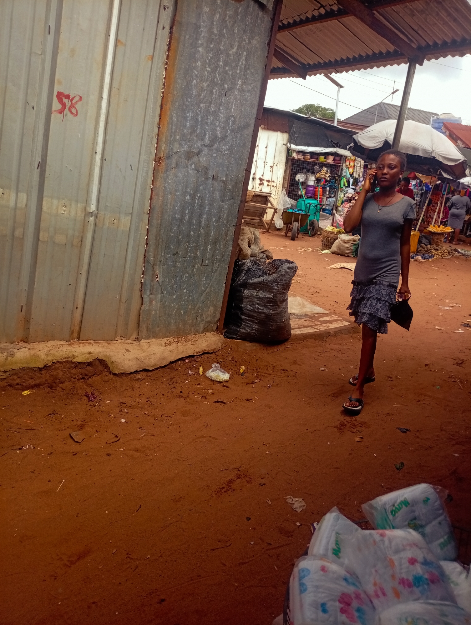 A young woman is walking and talking on her cell phone in a bustling outdoor market. She is wearing a gray t-shirt and a blue patterned skirt. She carries a black clutch in her right hand. The market appears to be selling various goods, including food items such as corn, baskets of produce, and what appear to be diapers packaged in plastic. In the background, there are stalls with shelves displaying goods and a woman wearing a gray dress. The ground is covered in reddish-brown dirt, and there is some scattered debris. A large, black plastic bag or sack is visible on the left side of the frame, next to a corrugated metal wall with red markings that read "58". The scene is under a covered roof structure, suggesting it might be a market with temporary shelters. The overall atmosphere seems active and transactional. The lighting suggests it is daytime, possibly overcast. The location cues of the market setting and the reddish soil are consistent with a place like Nkpor, Nigeria.