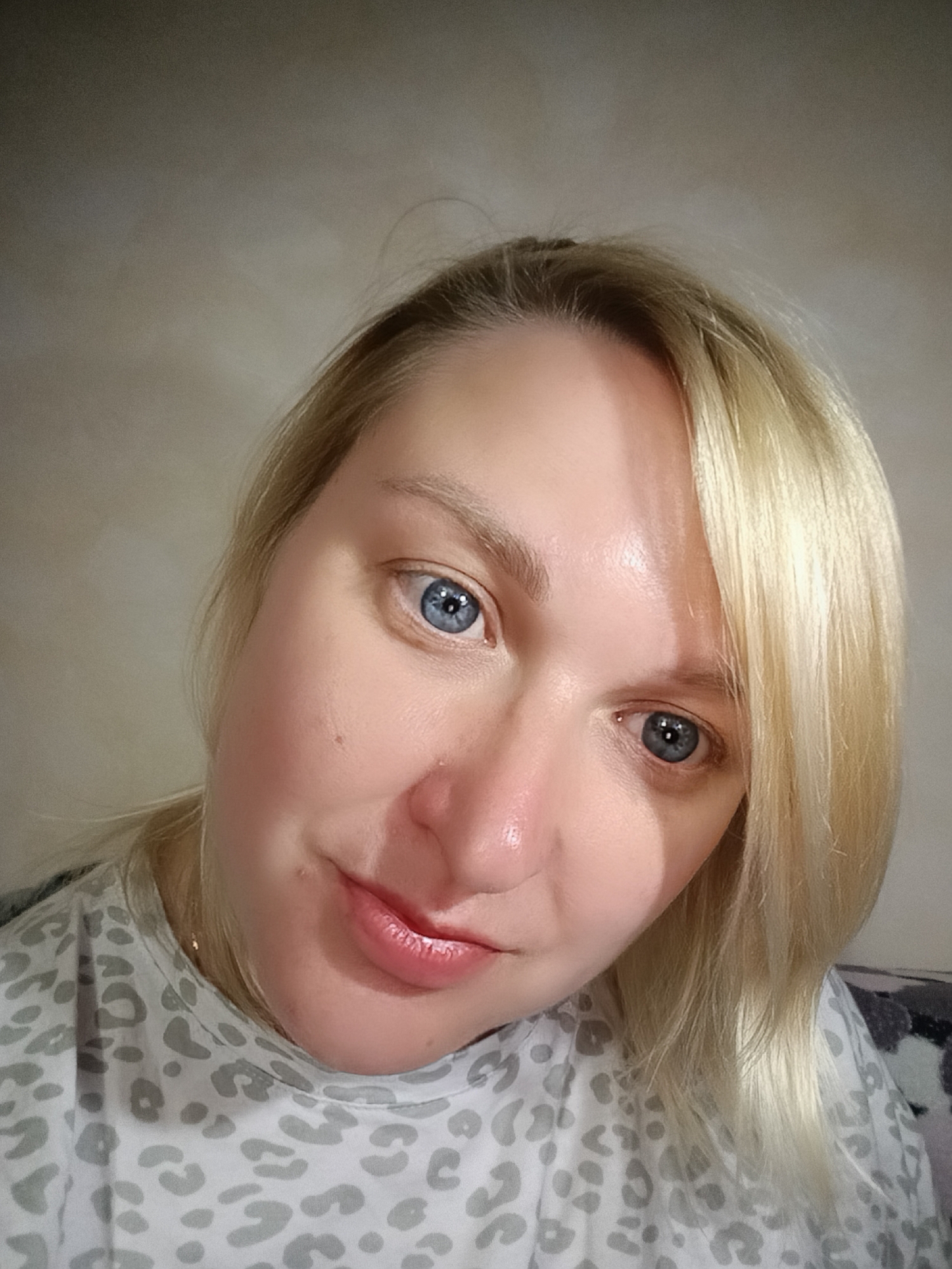 A fair-skinned woman with light blonde hair and blue eyes is the primary subject of the image. She is looking directly at the camera with a slight smile. Her skin appears smooth, with some shine visible on her forehead and nose. She is wearing a light-colored top with a gray animal print pattern. The background is a plain, light-colored wall. There is no indication of the location being in Chaikovsky, Russia. The image focuses solely on the woman's face and upper body. No specific actions or interactions are occurring in the image.