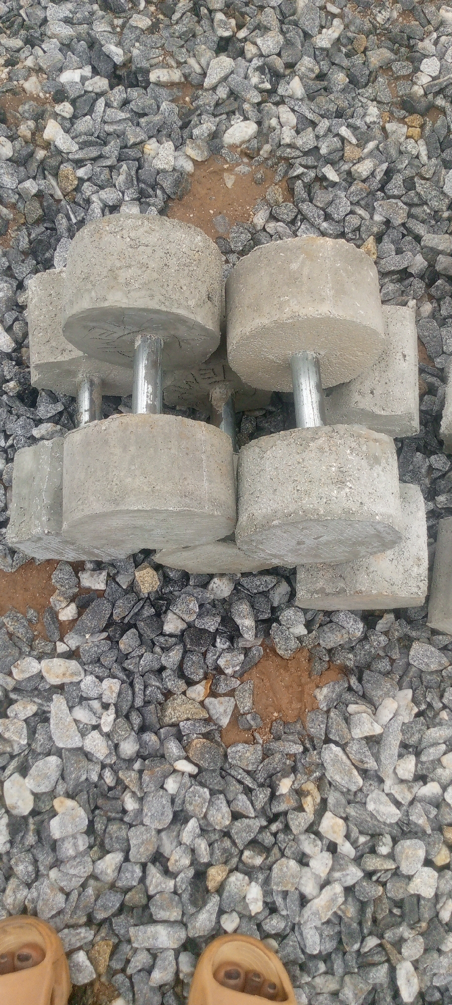 The image shows a pile of concrete dumbbells with metal handles, resting on a bed of gravel and sand. The dumbbells are arranged in two rows, with three in the front and three in the back, partially obscured. The gravel is a mix of gray, white, and brown stones, with some patches of damp brown soil visible. The overall scene appears to be an outdoor storage or construction area. The location of Indo, Nigeria is not explicitly depicted in the image, and there are no people or identifiable landmarks present.