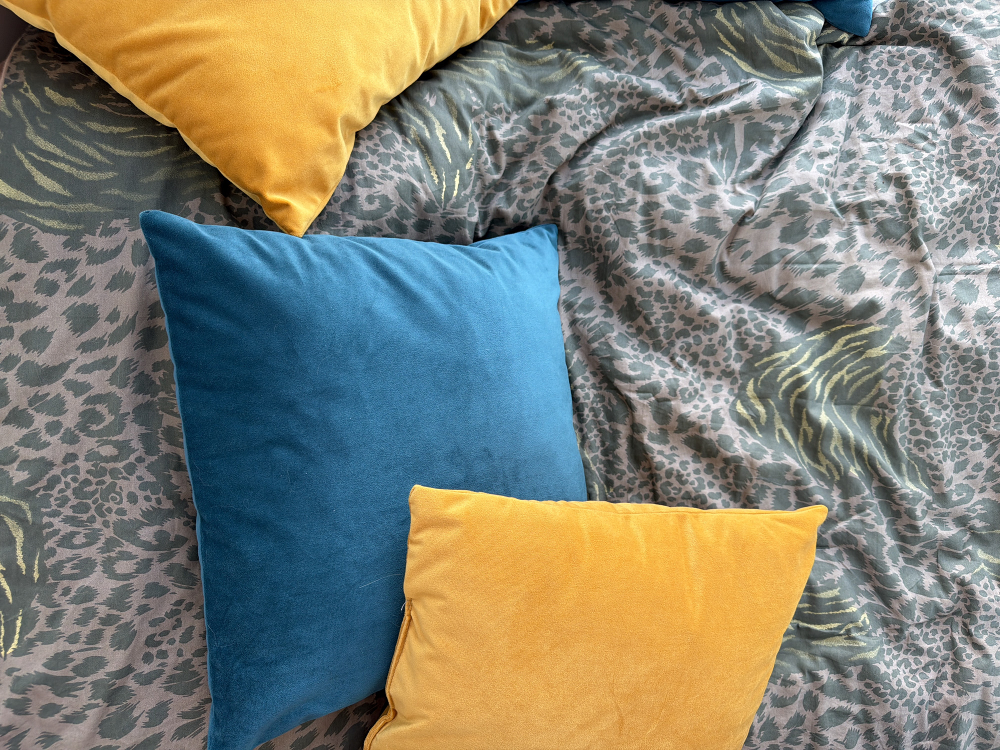 The image shows a close-up, top-down view of several decorative pillows arranged on patterned bedding. The bedding has a mixed animal print design featuring both leopard and zebra patterns in muted green and grey tones with subtle gold accents. Three pillows are visible: two are yellow velvet and one is blue velvet. The blue pillow is square and positioned in the center, with one yellow pillow partially underneath it and the other yellow pillow towards the top left corner. The arrangement appears to be indoors, likely in a bedroom setting.