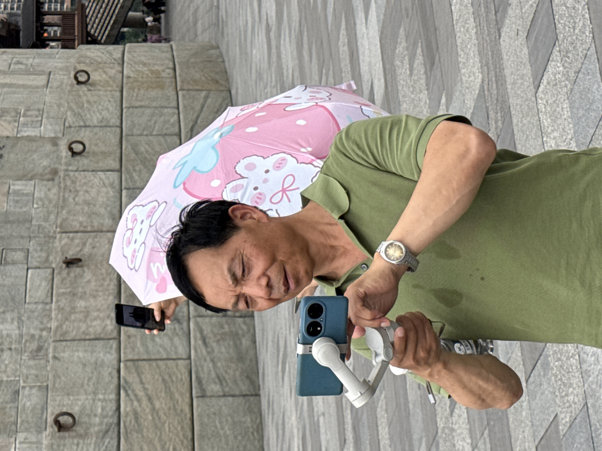 In Yuzhong District, China, a man in a green polo shirt is holding a smartphone on a gimbal stabilizer and appears to be recording a video. He is wearing a wristwatch on his left wrist. Behind him, a person is holding up a pink umbrella with cartoon bunny and strawberry designs. The background features a stone wall and paved ground.