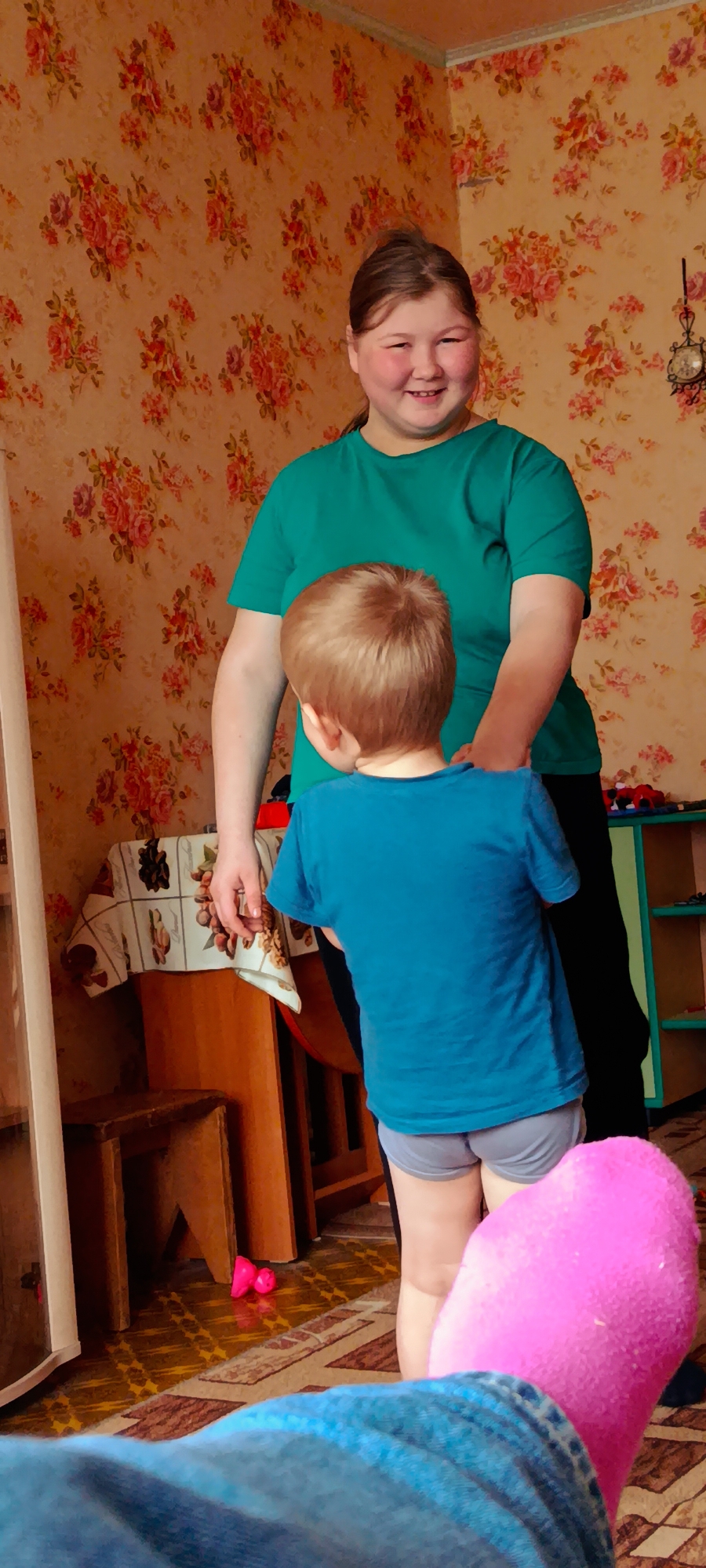 Here is a description of the image:
Two people are inside a building, likely a home. An adult woman with fair skin is standing and smiling, wearing a green shirt and black pants. In front of her is a young child with blonde hair and a blue shirt. The child is wearing underwear, and the back of their body is facing the camera. The woman appears to have her hands near the child's shoulders.
The interior has floral wallpaper. A table with a patterned tablecloth is visible to the left. Other furniture can be seen, including a mirror and a shelf unit with toys. A pink sock is prominent in the foreground, near the lower edge of the frame, suggesting someone is sitting. The location may be in the city of Фоки, Russia.