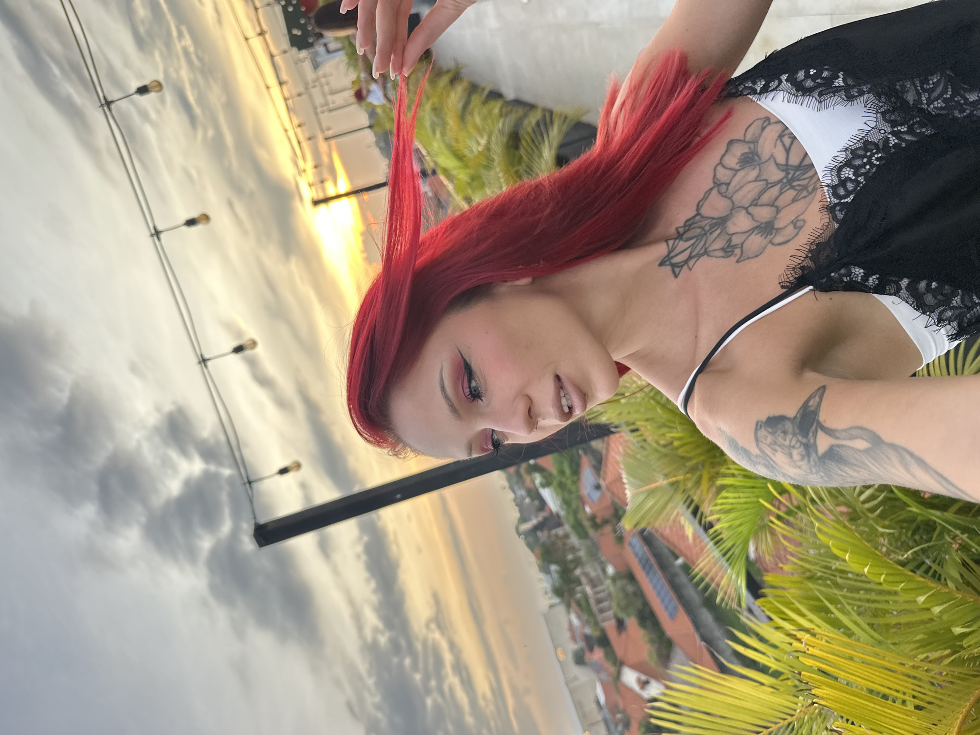 A woman with red hair stands on a rooftop in Tibubeneng, Indonesia, at sunset. She wears a black and white lace top. Visible tattoos are on her back and arm. She holds a strand of her hair. String lights are strung above. Palm trees grow on the rooftop. The cityscape is visible in the background. The sky is cloudy with golden hues from the setting sun.