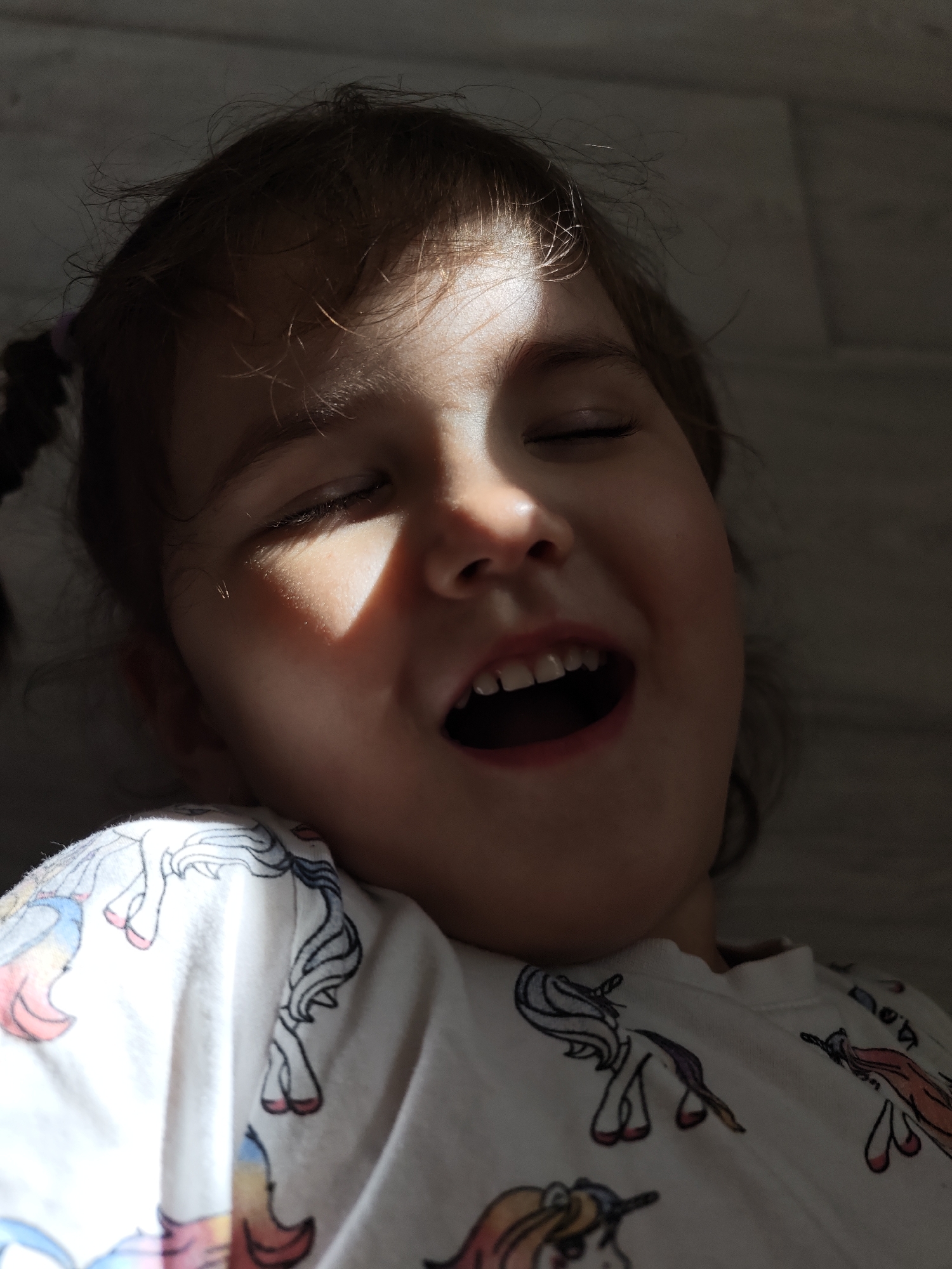 A close-up, low-angle shot depicts a young child with pigtails, eyes closed, and mouth open in a possible yell or laugh. Sunlight casts a harsh shadow across their face. The child is wearing a white shirt with a unicorn pattern. The background is a blurry gray wall. There is no indication this photo was taken in Уральское, Russia.