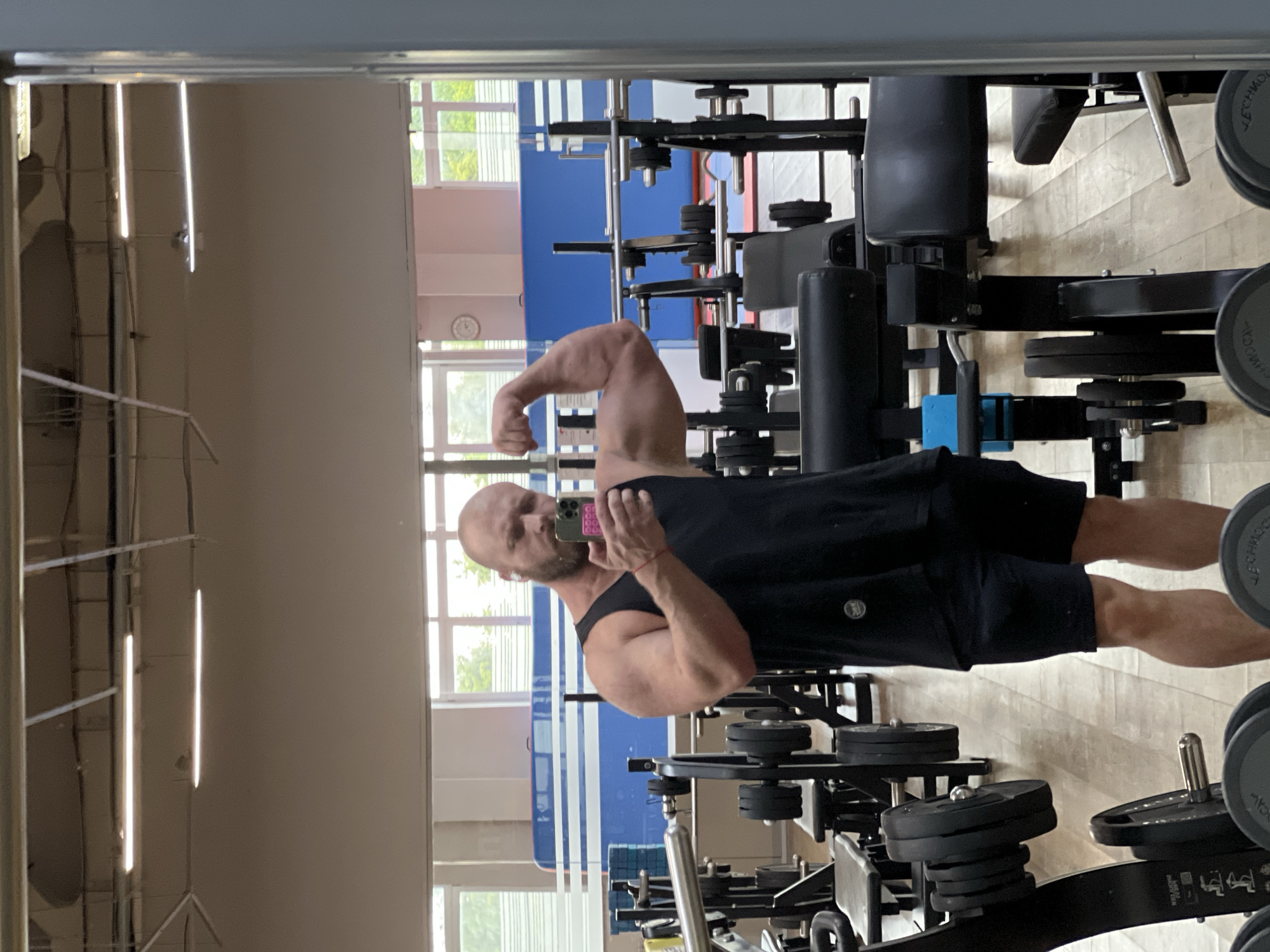 A bald, bearded man in a black tank top and dark shorts is taking a selfie in a gym. He is flexing his bicep. The gym has wooden floors, various weightlifting equipment, and large windows. The image appears to be taken in the city of Chaikovsky, Russia.