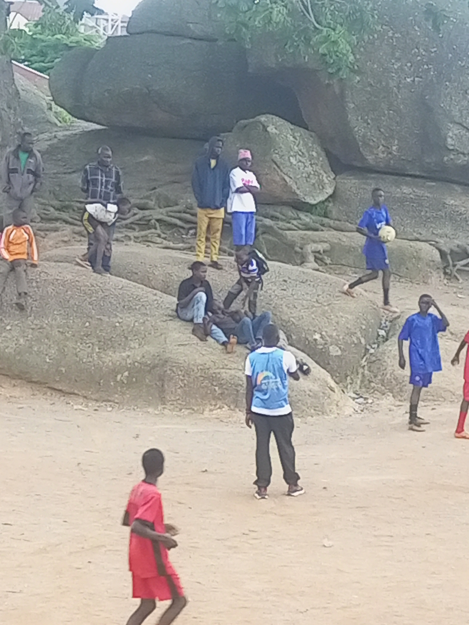 The image captures a group of individuals, likely children and young adults, gathered on a rocky, sandy terrain with large boulders in the background, characteristic of the landscape around Jos, Nigeria. Several figures are engaged in what appears to be a casual game of soccer, with one person holding a soccer ball. Others are standing, sitting, or walking around. The attire of the individuals varies, with some wearing casual clothing like t-shirts, shorts, and pants, while a few are in what could be soccer uniforms. The environment suggests an outdoor recreational area, possibly a park or open space near rock formations. The overall impression is of a community gathering, with the children and young adults interacting and enjoying an activity together.