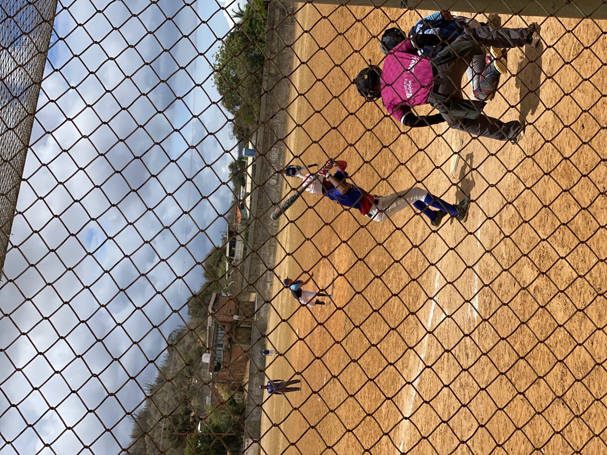 The image shows a baseball game in Altagracia, Venezuela, viewed through a chain-link fence. A batter in a blue and red uniform swings at a pitch. A catcher in a pink shirt and protective gear is positioned behind home plate. Other players are visible on the field. The field is dirt. Buildings and trees are visible in the background. The sky is cloudy.