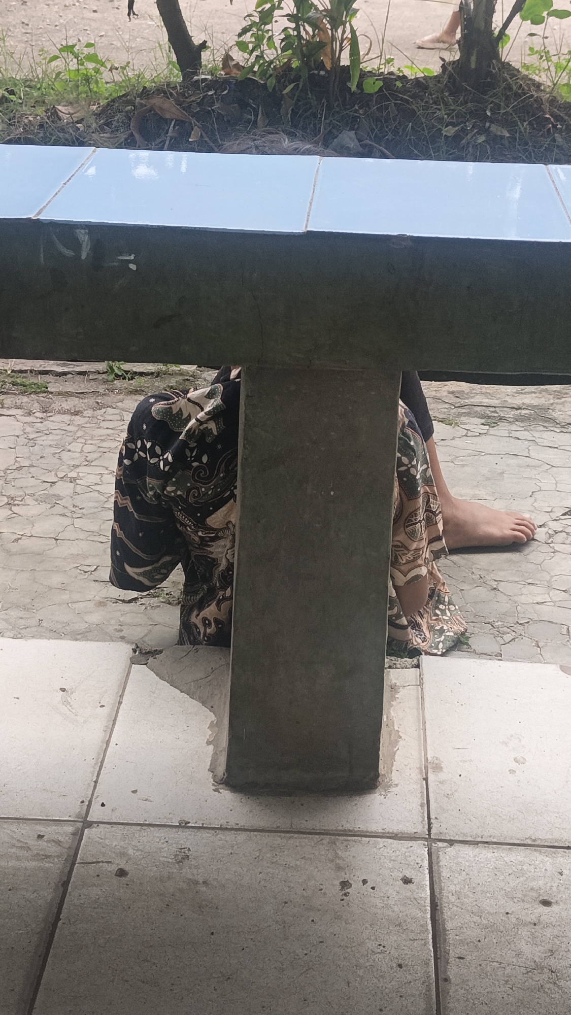 A person wearing a patterned skirt is sitting on the ground in Tigaraksa, Indonesia. They are partially hidden behind a concrete pillar that is supporting a tiled structure. The ground is paved with square tiles, and there is vegetation visible behind the tiled structure. The person's bare foot is visible on the right side of the image.