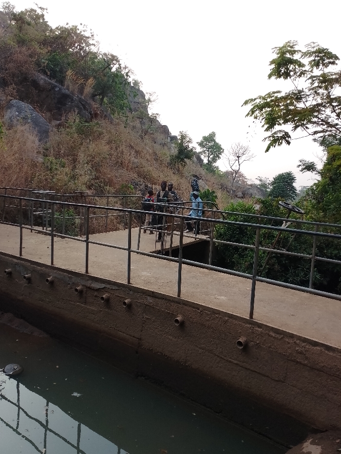 The image captures an outdoor scene featuring a combination of natural landscape and man-made structures, with a small group of people in the midground.
In the foreground, a concrete channel filled with murky, greenish water occupies the bottom left portion of the frame. The inner walls of this channel are coated with a thick layer of dark, textured grime or sediment. Protruding horizontally from the upper part of the channel's inner wall are several short, round pipe sections, evenly spaced, suggesting drainage outlets or inlets. A reflection of the metal handrail above is visible on the surface of the water.
Immediately above the channel, running horizontally across the midground, is a concrete pathway or bridge. A sturdy metal handrail, consisting of vertical posts and horizontal bars, lines the pathway on both sides, though the right side railing extends downwards towards what appears to be a lower area.
A small group of approximately five to six individuals is gathered on this pathway, positioned towards the right side of the frame. One person, wearing a light blue long-sleeved shirt, stands facing towards the right. Next to them, another individual in dark clothing, possibly a dark blue or black uniform, stands. Further to the left within the group, other individuals are visible, including what appears to be a child wearing a red and white striped top. Their faces are not clearly discernible due to the distance and resolution, but they appear to be standing and observing.
Beyond the pathway and the people, the landscape consists of a steep, rocky hill rising prominently in the background on the left and central parts of the image. This hillside is largely covered in dry, light brown or yellowish scrub and sparse vegetation, with numerous large, grey rock formations exposed. Some scattered trees, appearing somewhat dry or leafless, are also present on the hill.
To the far right in the midground, below the pathway, there is a dense cluster of green foliage and trees, indicating a more lush vegetated area, possibly a valley or lower ground. A distinct, large, dark-colored wheel-like mechanism, possibly part of a valve or machinery, is visible amidst this green vegetation. The sky above is bright and almost white, suggesting either an overcast day or the bright conditions of late morning or early afternoon. No discernible text is visible in the image.