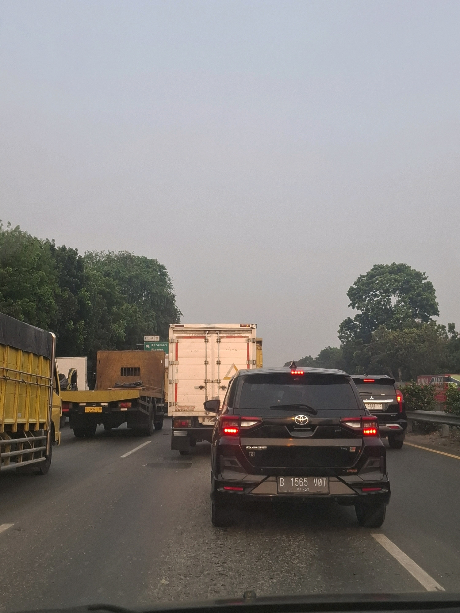 The image depicts a traffic jam on a highway in Kelapa Dua, Indonesia. Several vehicles, including trucks and cars, are stationary on the asphalt road under an overcast sky. Trees line the sides of the highway. A green signpost with white text indicating "KELUAR 21 Karawaci" is visible. The vehicles include a yellow truck on the left, a white box truck in the middle, and several cars, one of which is a black SUV with the license plate B 1565 V0T. The environment suggests a typical urban commuting scene.