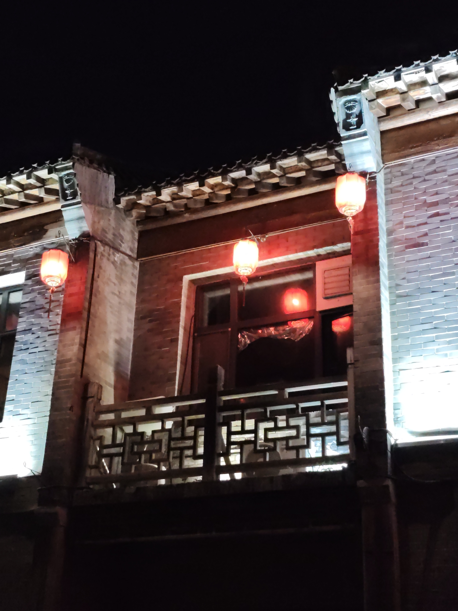 The image shows a facade of a traditional brick building at night, illuminated by red lanterns. The building has a tiled roof with ornate eaves and decorative corner elements. There is a balcony with a latticework railing. Windows with dark frames are visible, and reflections of lights can be seen in the glass. The lower part of the building is dark, suggesting it is at street level or below. The overall impression is one of an atmospheric, historical streetscape. The city of Lindian, China, is mentioned as the location, implying this scene is within a historically preserved area or a district with traditional architecture. There are no people visible in the image.