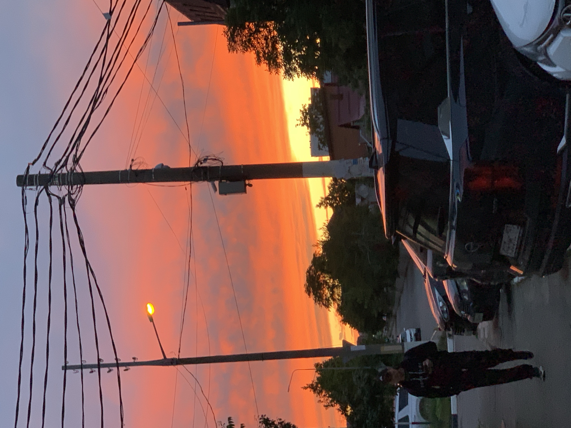 The image captures an outdoor urban or suburban street scene at either sunset or sunrise, dominated by a vibrant, multi-hued sky. The horizon is ablaze with intense orange and yellow light, which gradually transitions into softer pinks, purples, and light grey-blue towards the upper sky.
In the left foreground, a tall utility pole stands prominently, supporting a complex network of dark electrical wires that crisscross the frame. Further to the right, a smaller utility pole features a single, lit street light casting a warm yellow glow against the dramatic sky.
On the right side of the image, several dark-colored cars are parked along the street. The rear of a dark SUV or sedan is visible in the immediate right foreground, showing a license plate that is illegible due to distance and angle.
A person, dressed in dark clothing including a jacket and patterned pants, is standing on the street in the lower right portion of the frame. They appear to be looking towards the horizon or away from the camera. Scattered trees with green foliage are visible in the midground and background, partially obscuring distant buildings. The street surface is dark, suggesting asphalt or pavement.