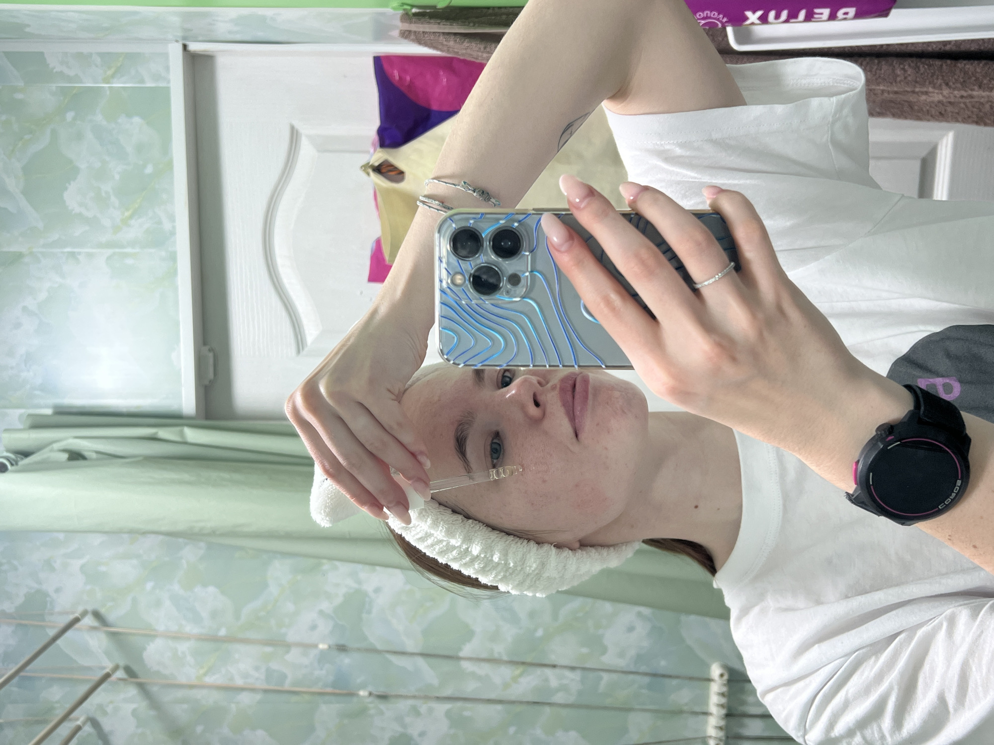 A woman is taking a selfie while applying serum to her face. She is wearing a white headband and a white t-shirt. She is holding a silver iPhone with a blue-and-silver patterned case. She has a silver ring on her finger and a black smartwatch on her wrist. She is in a bathroom with green and white marble walls and a white cabinet. In the background, there is a towel rack and a box labeled "BETOX". The location is likely a bathroom in a residential building in the city of Chaikovsky, Russia, as the user has provided this information.