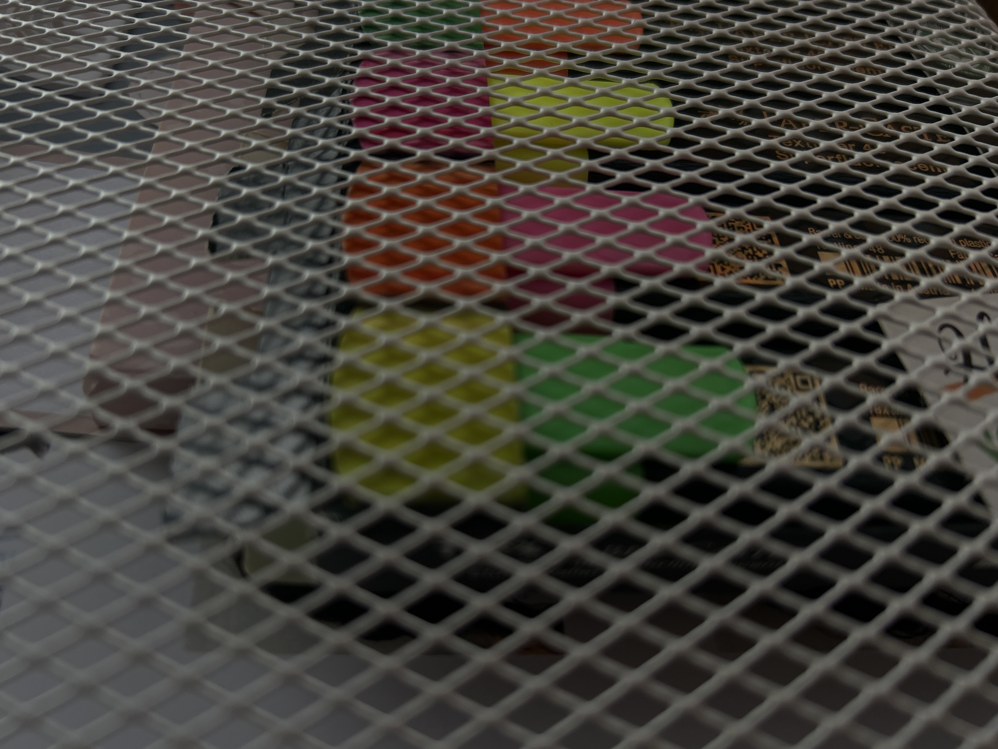 The image shows a metal wire mesh in the foreground, obscuring the view of items behind it. The items appear to be stacked containers of various colors including pink, orange, yellow, and green. The wire mesh is close to the camera and out of focus in some areas. Some printed materials with text and QR codes are also visible behind the mesh. The image location is not apparent.