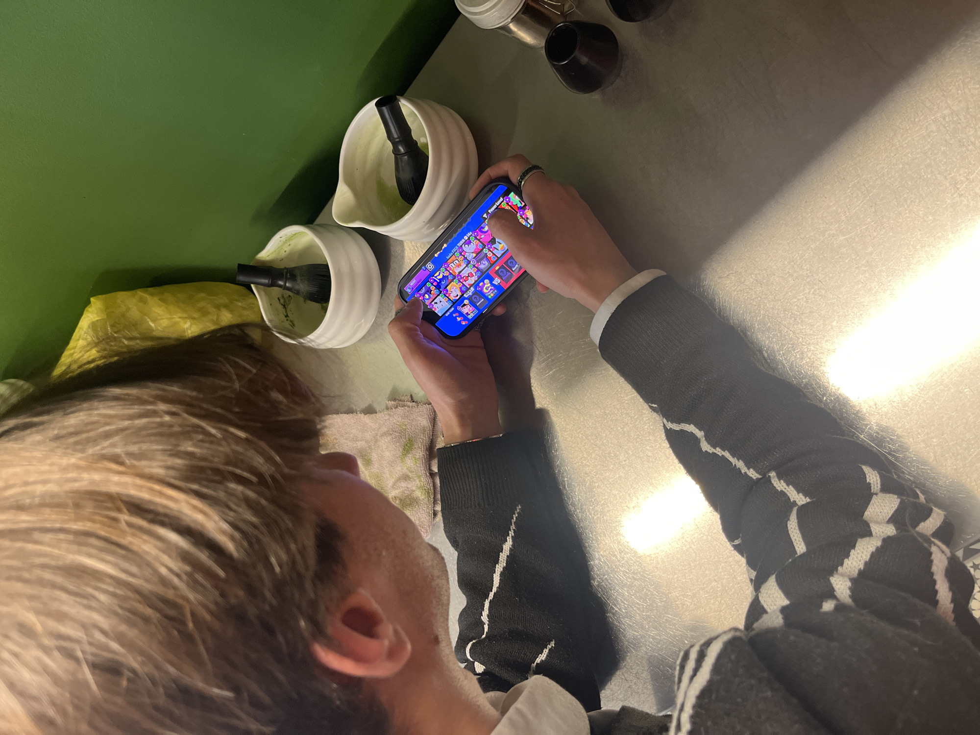 A person is seen from an overhead perspective, playing a game on a smartphone. The person has light brown hair and is wearing a black and white striped sweater. The hands are visible, holding and manipulating the phone. The phone screen displays a brightly colored puzzle game. To the left, there are two white bowls containing black utensils, possibly for preparing a beverage. Behind the bowls is a green surface, possibly a wall. The scene is illuminated by light coming from the right side of the frame. The location is unknown, and there is no information about Sofia, Bulgaria in the image.