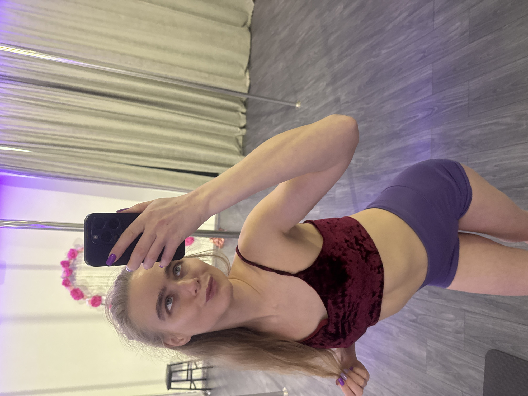 A full-body selfie of a young woman is depicted, taken indoors and rotated 90 degrees clockwise. The woman, with fair skin, long blonde hair, and light-colored eyes, is the central subject. She is dressed in a maroon velvet crop top with thin straps and purple athletic shorts. Her fingernails are painted purple.
She holds a black smartphone with a multi-lens camera in her right hand, taking the picture herself. She looks directly at the camera with a subtle, confident smile. Her left arm is bent, and her left hand lightly touches a strand of her hair.
The setting appears to be a fitness or dance studio. To her left, a shiny metal pole, commonly used for pole fitness, extends vertically. The floor is covered with grey-toned wood laminate. In the background, there are light-colored curtains or blinds, and decorative elements include a white circular frame adorned with pink floral accents. Purple accent lighting is visible along the left edge of the frame. The overall atmosphere suggests a casual moment during or after a workout session. No text is visible in the image.