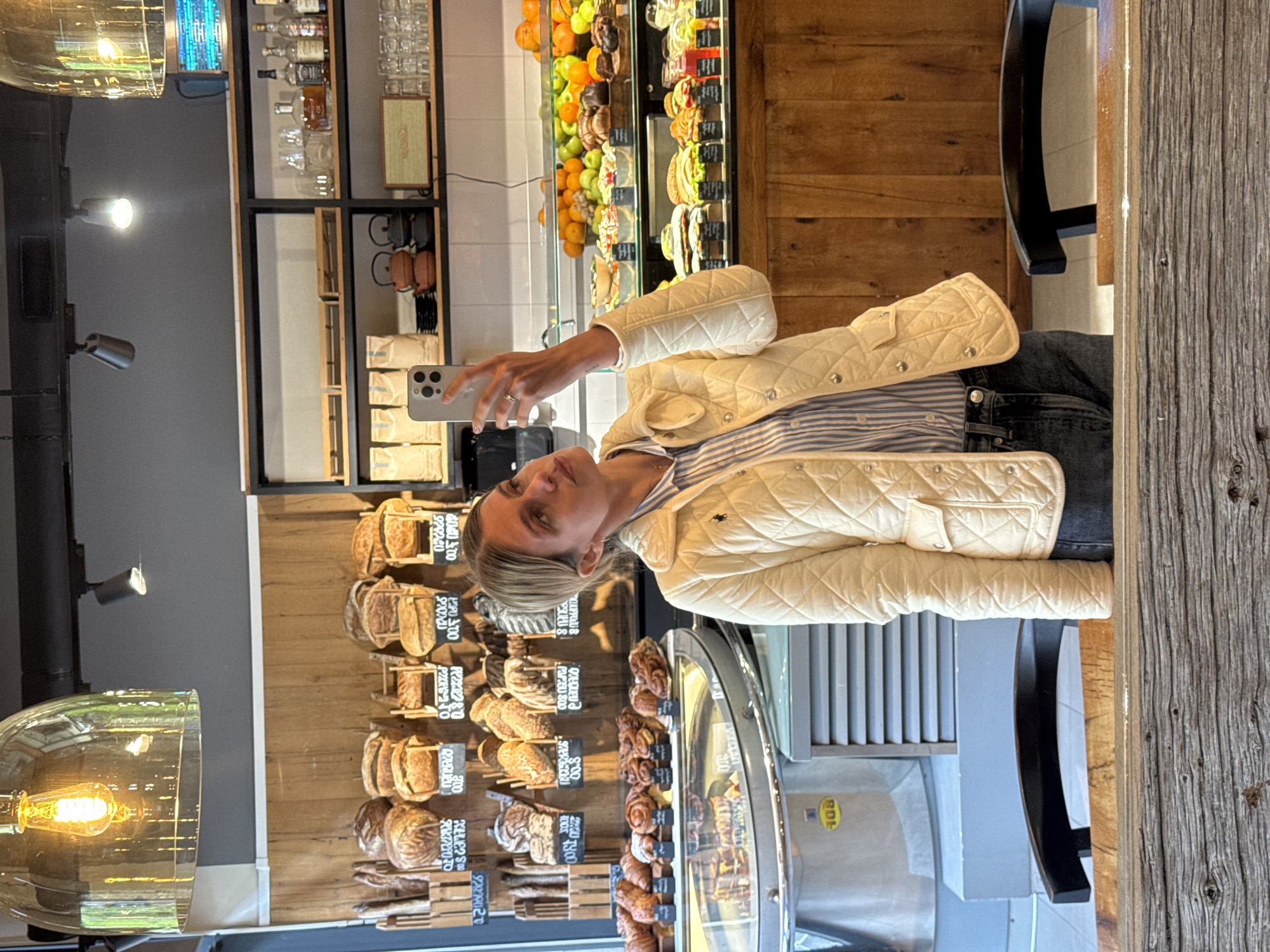 An image, rotated 90 degrees clockwise, captures a woman taking a selfie in what appears to be a modern bakery or cafe in Tbilisi, Georgia. The woman, with light-colored hair, is dressed in a blue and white striped shirt layered under a cream-colored quilted jacket and jeans. She holds a silver-colored smartphone, likely an iPhone, in her right hand, directed towards her face to capture the image.
The setting is an indoor space characterized by wooden elements and glass display cases. To the left, various loaves of bread and baguettes are neatly arranged on wooden shelves against a textured wooden wall. In the center, below the woman, a refrigerated display case showcases an assortment of pastries, including cinnamon rolls and other baked goods. Further in the background, another glass display case holds fresh fruits like oranges and green apples. On a shelf above these displays, bottles of various spirits, glassware, and a wooden speaker are visible, suggesting a bar or beverage section.
The cafe features stylish lighting, including a large glass pendant light with an Edison bulb in the lower left foreground, and several smaller spotlights. Part of a rustic wooden table and a dark chair are visible on the right.
Visible text on the bread labels and displays includes numbers and words, some of which appear to be in Georgian script, likely indicating prices or product names. Examples from OCR include "QUFFOCUS", "QUEU 8", "D", "SOO", "Оян", "허용" (a Korean word), and numerical values such as "00.8", "20.00", "5.00", "9.00", "13.00", "2.00", and "4.2060826". The overall ambiance suggests a well-lit, inviting establishment.