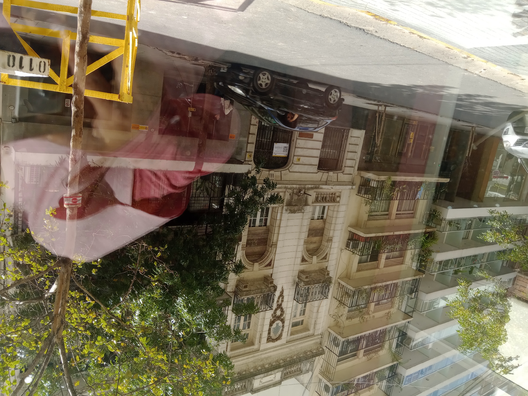 The image shows a reflection of a street scene in Buenos Aires, Argentina. A black car is parked on the street, with buildings lining the road. The architecture includes both older, ornate structures and more modern, sleek buildings. A yellow barrier is visible on the left side of the frame, partially obscuring the view. A tree is also visible, its branches and leaves intermingling with the reflections. The reflections are distorted, indicating the presence of glass or a reflective surface. A stop sign is reflected.