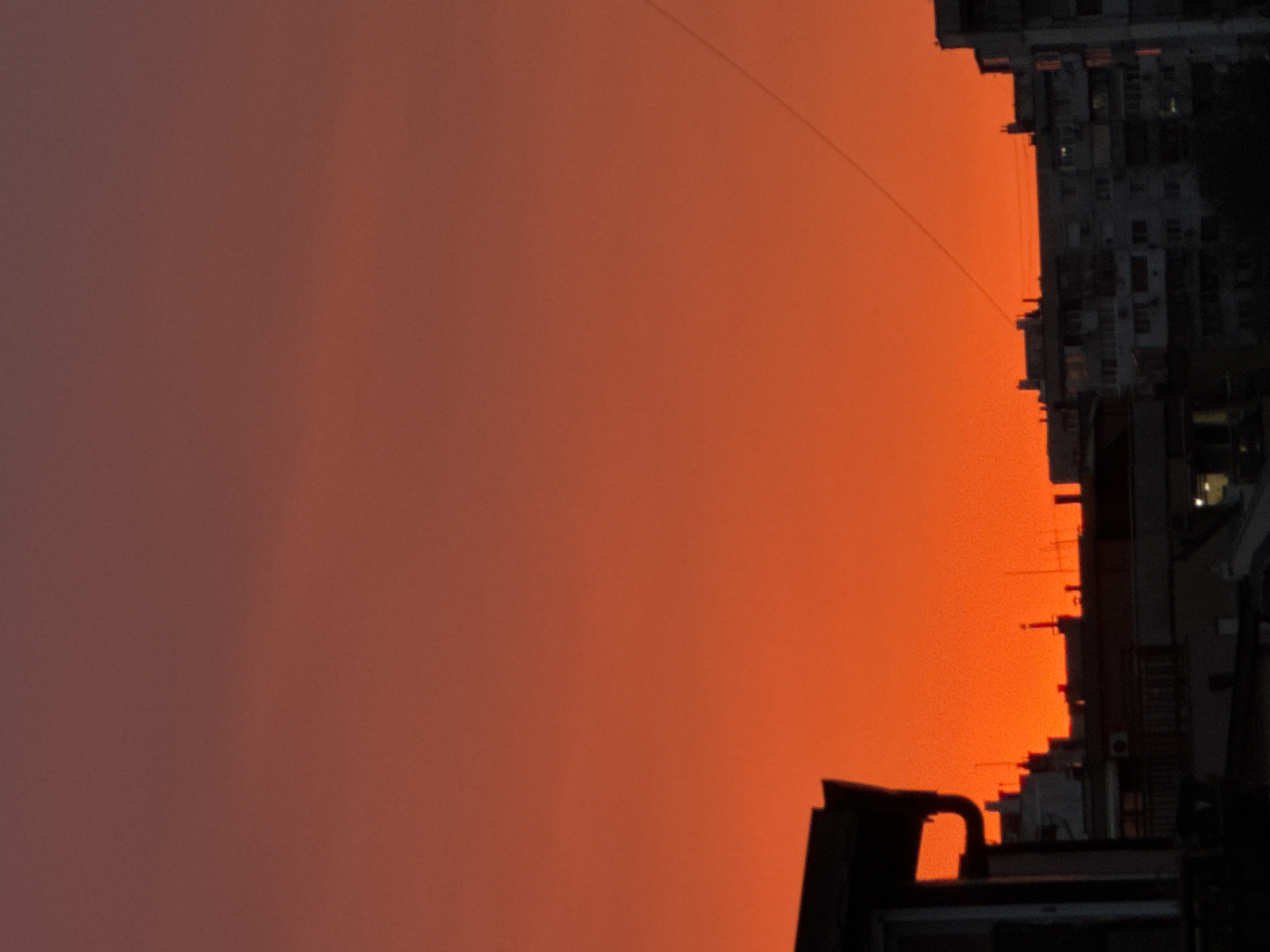 The image depicts a cityscape silhouetted against a vivid orange sky, likely during sunset or sunrise. The structures on the right side of the image appear to be buildings, possibly residential or commercial, densely packed together. A power line is faintly visible crossing the sky. The dark objects in the foreground at the bottom are unclear but could be building elements or rooftops. The specific location in Varna, Bulgaria, cannot be identified based on the provided image alone.
