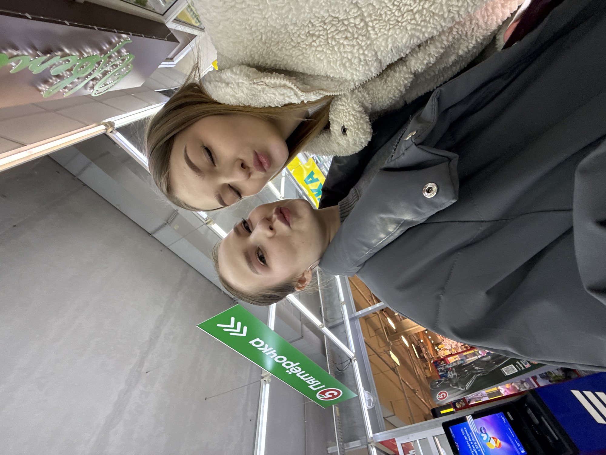 Two young women pose together in what appears to be the "Pyaterochka" supermarket. The supermarket's green and white logo is visible in the foreground. The woman on the left is wearing a white fleece jacket, while the one on the right is wearing a gray jacket. The store's interior, including shelves and a self-checkout kiosk, is partially visible in the background. The image was likely taken in the city of Chaikovsky, Russia, based on the store's name and logo.