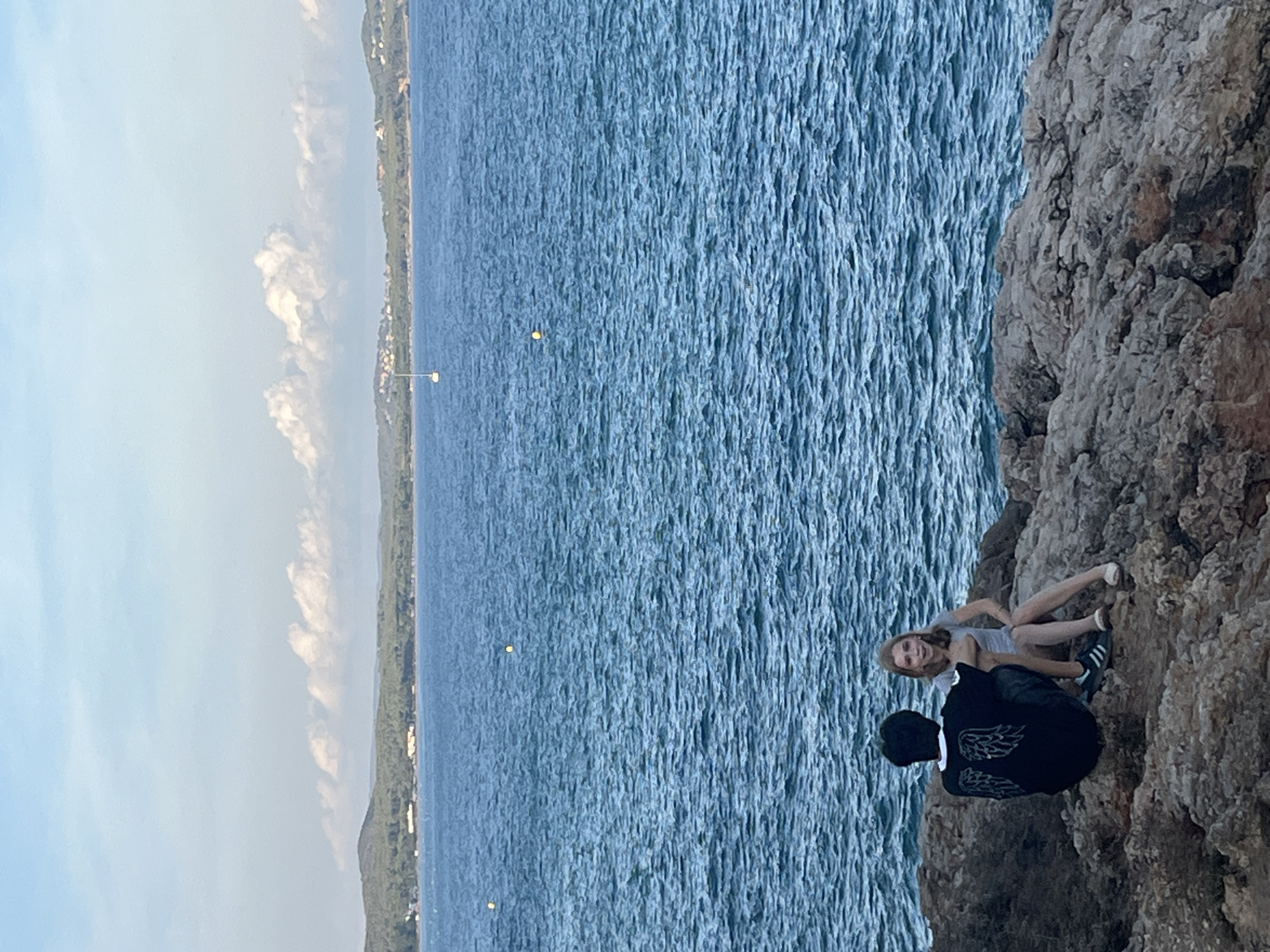 Two people are sitting on rocks by the sea. The person on the left is wearing a black t-shirt with white wings on the back and dark pants. The person on the right is wearing a light-colored t-shirt and light-colored pants. They are facing the water. The sea is a deep blue with small waves. In the distance, there is a coastline with trees and some buildings, and a few white clouds are in the sky. The location is the city of El Miracle, Spain.