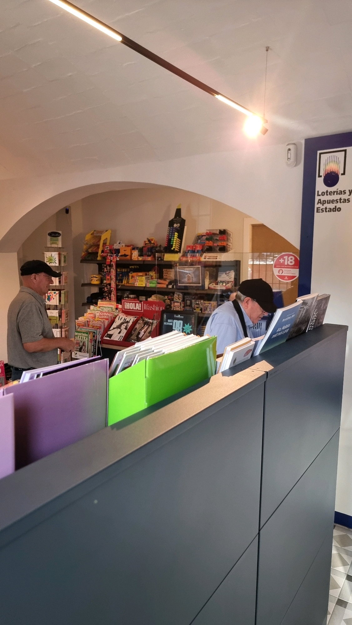 The image depicts the interior of a shop, likely a convenience store or newsstand, in Sant Joan Les Fonts, Spain. In the foreground, a dark gray counter with a metallic trim separates the viewer from the interior. On top of the counter are several magazine holders, some filled with magazines and others with blank white papers. Two prominent holders are visible, one filled with purple and one with bright green paper.
Behind the counter, a man wearing a gray shirt and a black cap stands on the left, looking at items on a shelf. He appears to be an employee or a customer. On the right, another man, also wearing a black cap and a light blue shirt, is bent over a counter, examining magazines. He also appears to be an employee.
The shelves behind them are stocked with various items, including magazines, newspapers, lottery tickets, packaged goods, and stationery. A prominent sign on the right wall advertises "Loterías y Apuestas del Estado." A circular sign with "+18" is also visible on a glass partition. The ceiling has an arched shape and is illuminated by a long, modern light fixture. The floor has a tiled pattern. The overall impression is a well-stocked retail environment.