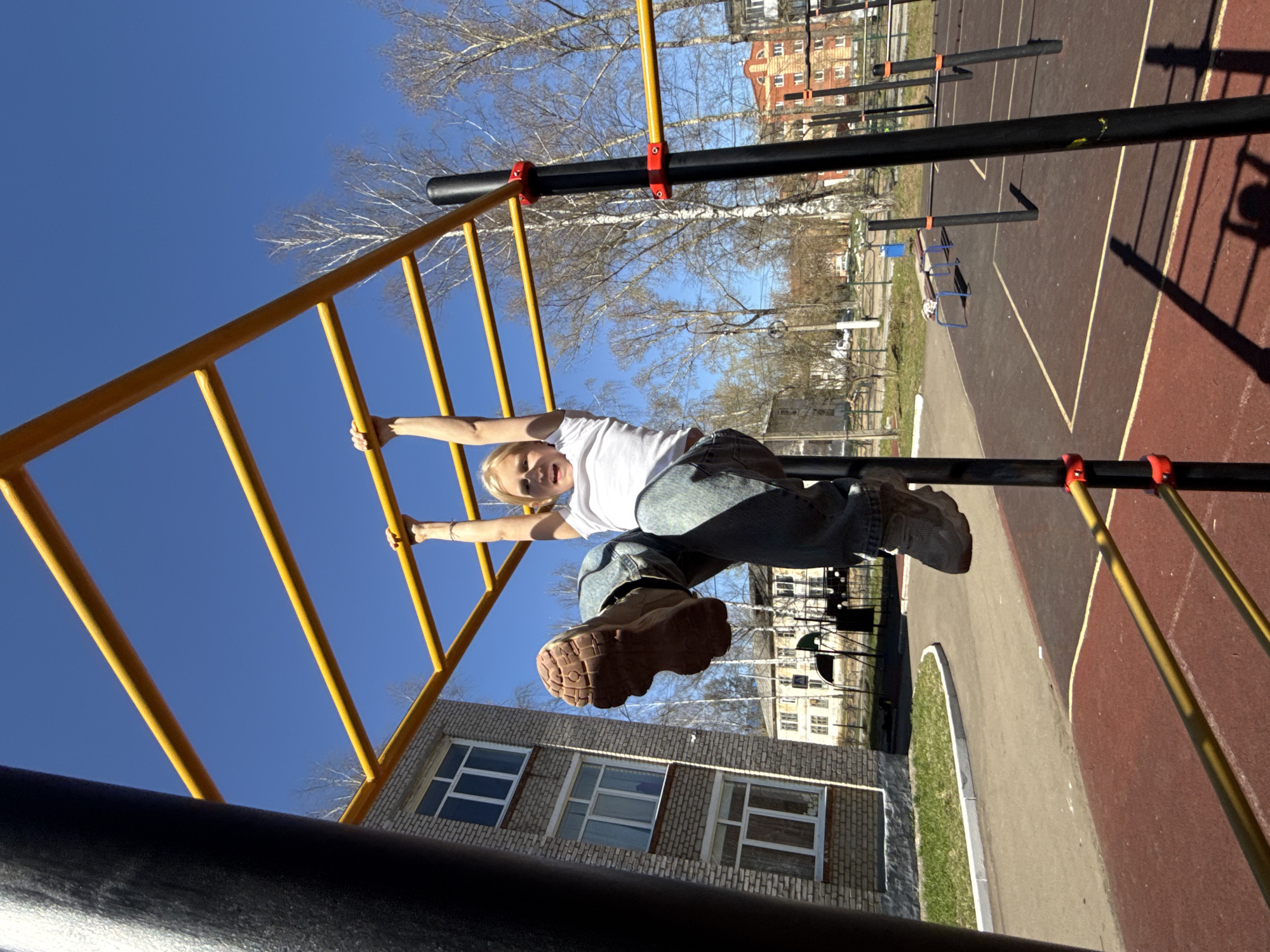 A low-angle, eye-level shot depicts a blonde child hanging upside down from yellow monkey bars in a playground in Chaikovsky, Russia. The child, wearing a white t-shirt and jeans, looks towards the camera. Their left leg is extended towards the camera, exposing the sole of their brown boot. Behind the child are the monkey bars and other playground equipment. A brick building with several windows is visible in the background on the lower right. Leafless trees are visible behind the playground, and a clear blue sky dominates the upper portion of the image. Shadows are cast by the playground equipment onto the red and gray surface of the playground.