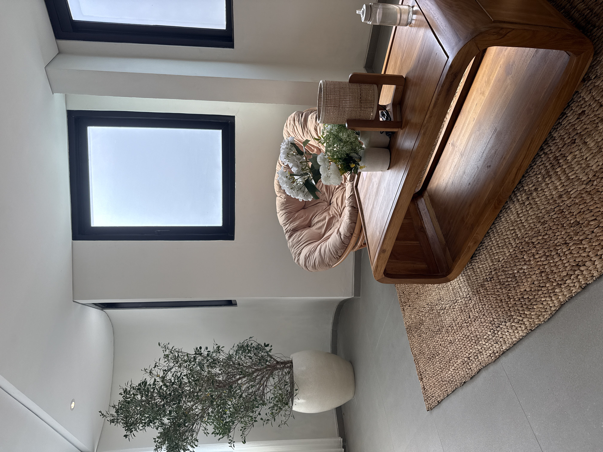 This media file captures a brightly lit, modern indoor living space, likely in Kuta Selatan, Indonesia. The image is oriented vertically, showcasing the room from a slightly elevated perspective.
In the mid-right, a distinctive wooden coffee table with a curved, sculptural design is prominent. Beneath it, and extending towards the bottom right, is a textured, natural fiber woven rug, possibly made of jute or seagrass. To the left of the coffee table, a light-colored, plush papasan-style chair with a ruffled cushion provides a soft seating option.
On the coffee table, a white vase holds an arrangement of artificial white flowers, resembling hydrangeas, alongside some delicate light green foliage, possibly baby's breath. A cylindrical woven lantern or decorative item and a clear glass carafe or water bottle are also present.
In the bottom left corner, a large, leafy potted plant, likely an olive tree, stands in a smooth, off-white, egg-shaped planter. The room features clean white walls, with architectural details including a horizontal beam visible between two black-framed windows at the top of the image. The flooring is a consistent neutral grey tile. A small, recessed light fixture can be observed on the wall towards the bottom left.
The natural light entering through the large windows appears diffused and bright, indicating daytime, possibly under an overcast sky or indirect sunlight. The overall aesthetic combines modern design with natural materials and a serene ambiance, suggesting a comfortable and inviting lounge or relaxation area. No activity is taking place, and no visible text is present in the image.